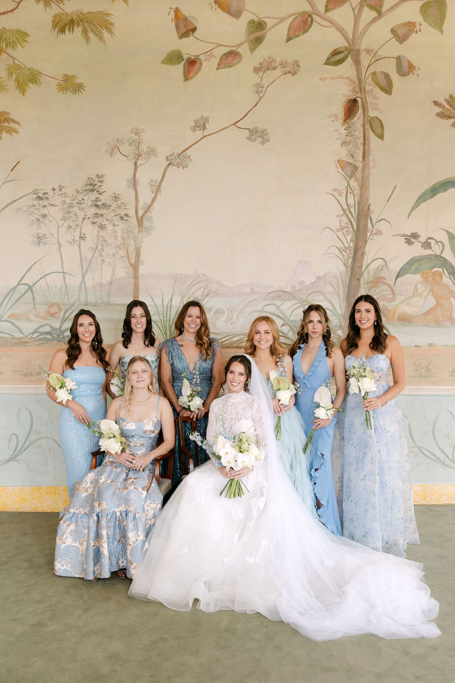 Bridesmaids in light blue floral dresses pose against a painted wall mural.