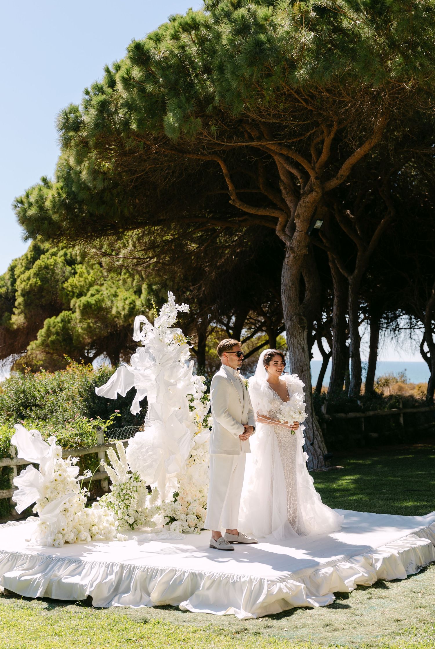 Outdoor wedding ceremony setup with white decor and floral arrangements under pine trees.