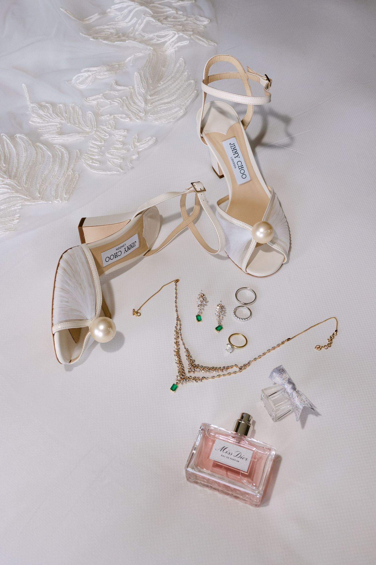 Wedding accessories including white heels, perfume bottle, and jewelry on light gray background.