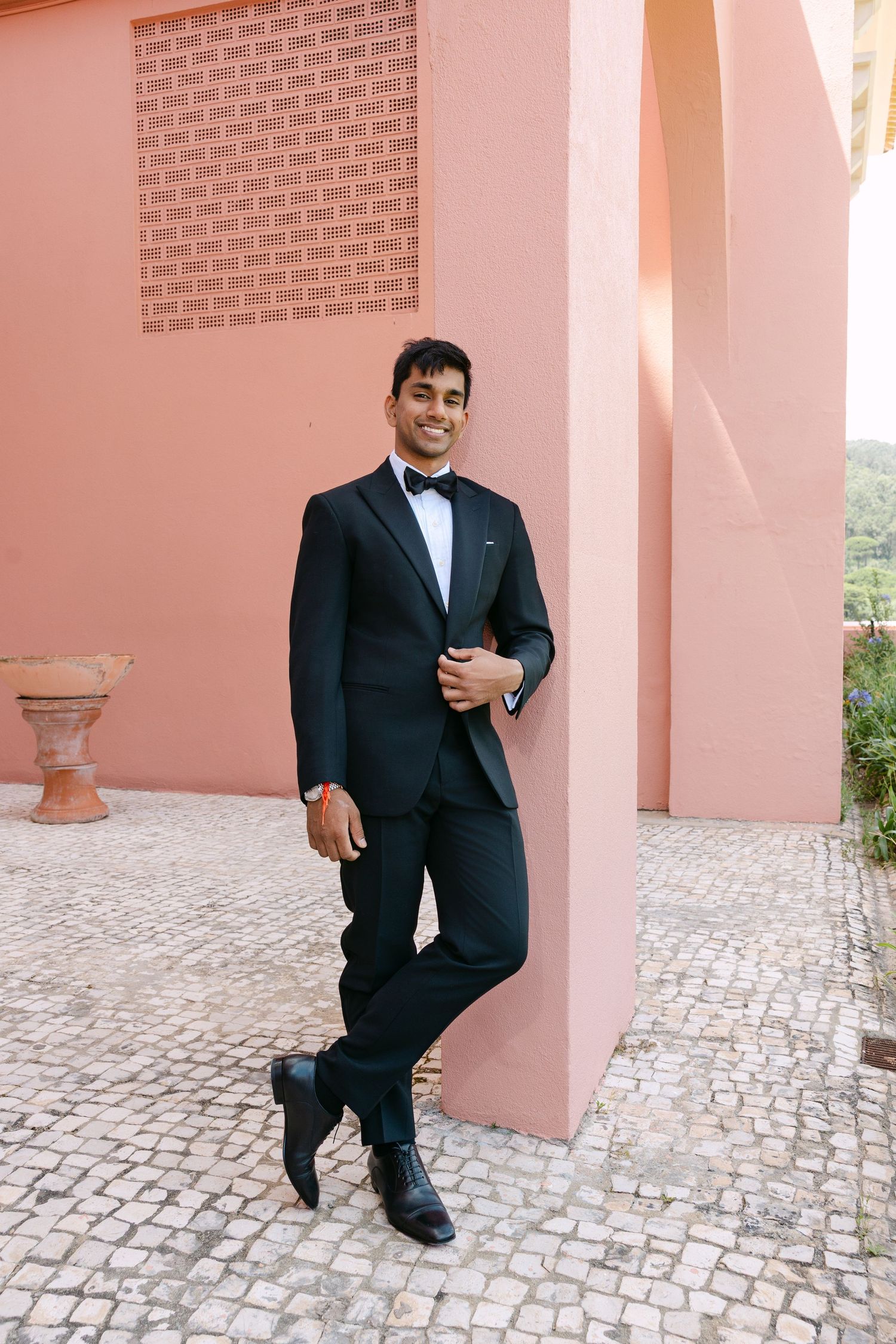 Groom in classic black tuxedo posing against pink stucco wall with cobblestone path.