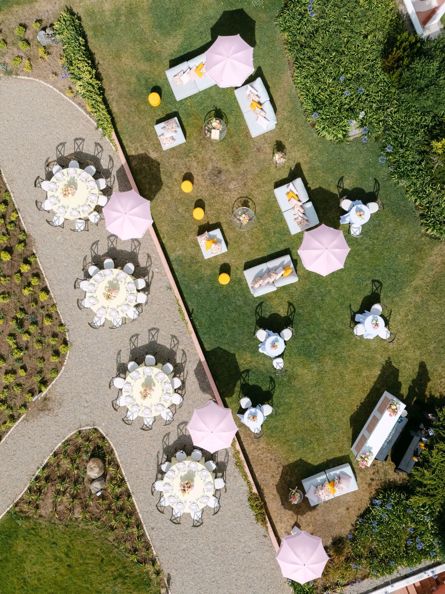 Aerial view of an outdoor wedding reception setup with round tables and pink umbrellas on grass and concrete.
