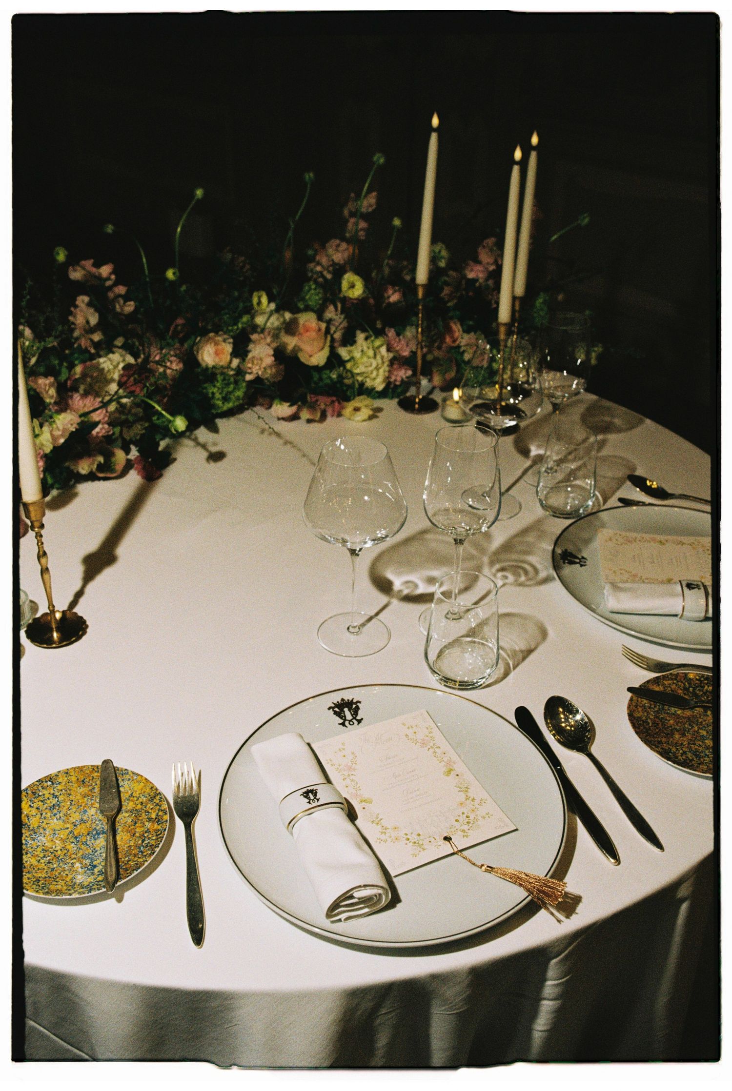 Elegant wedding reception table with white linen, crystal glasses, taper candles, floral centerpiece, and gold flatware.
