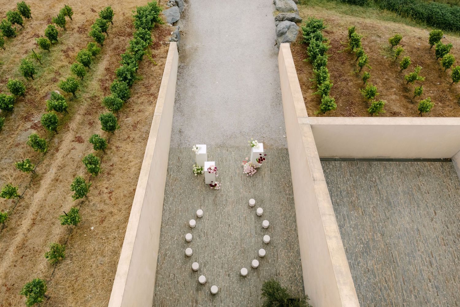 Aerial view of two people arranging white spheres in a heart shape on a stone path between vineyard rows.