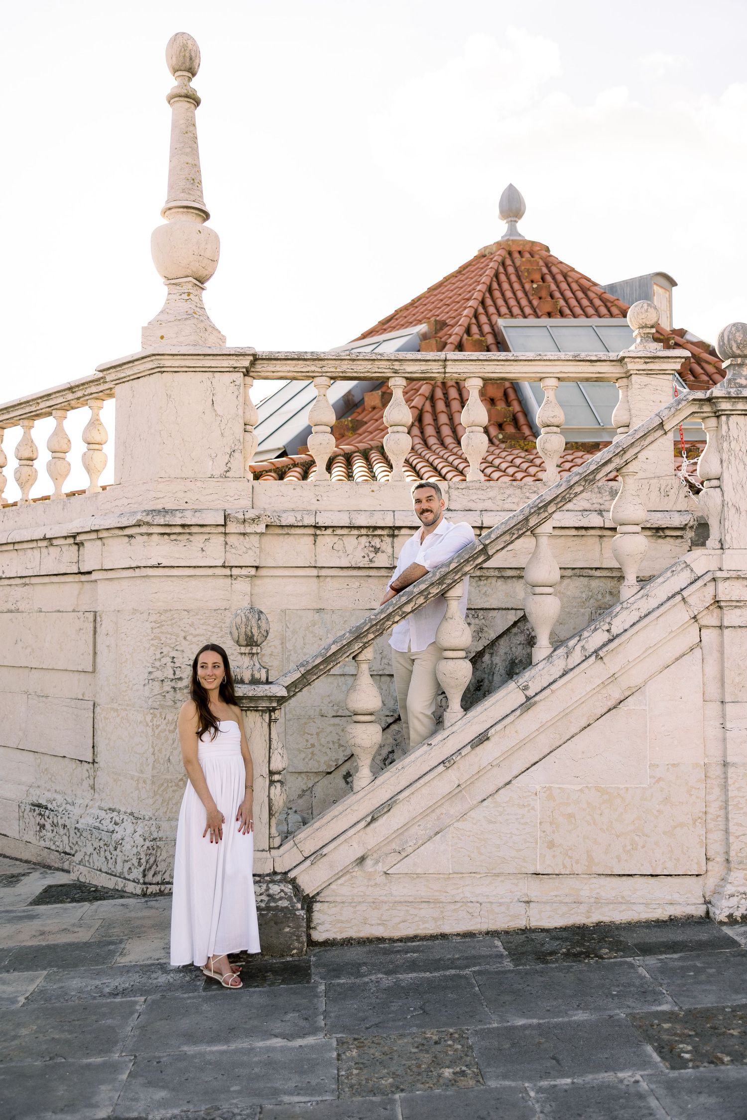 Couple in white outfits posing on historic stone staircase with ornate balustrade and terracotta roof building backdrop.