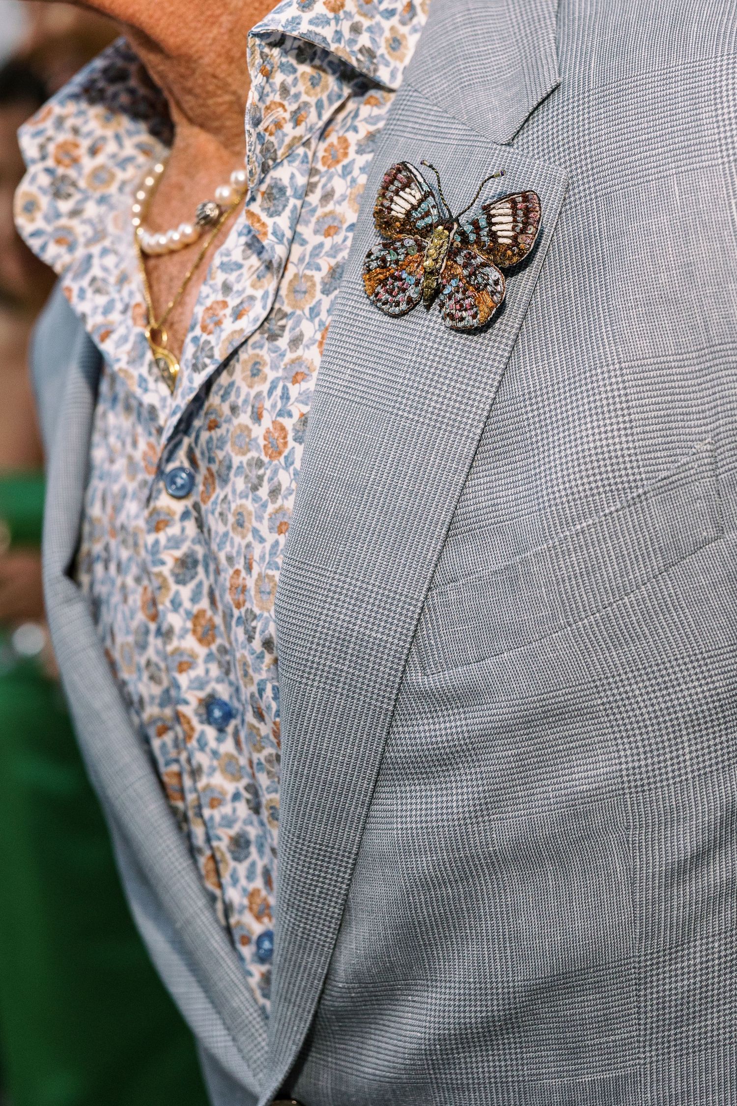 Close-up of a gray plaid blazer with a butterfly brooch on the lapel, worn over a floral shirt with pearl necklace.