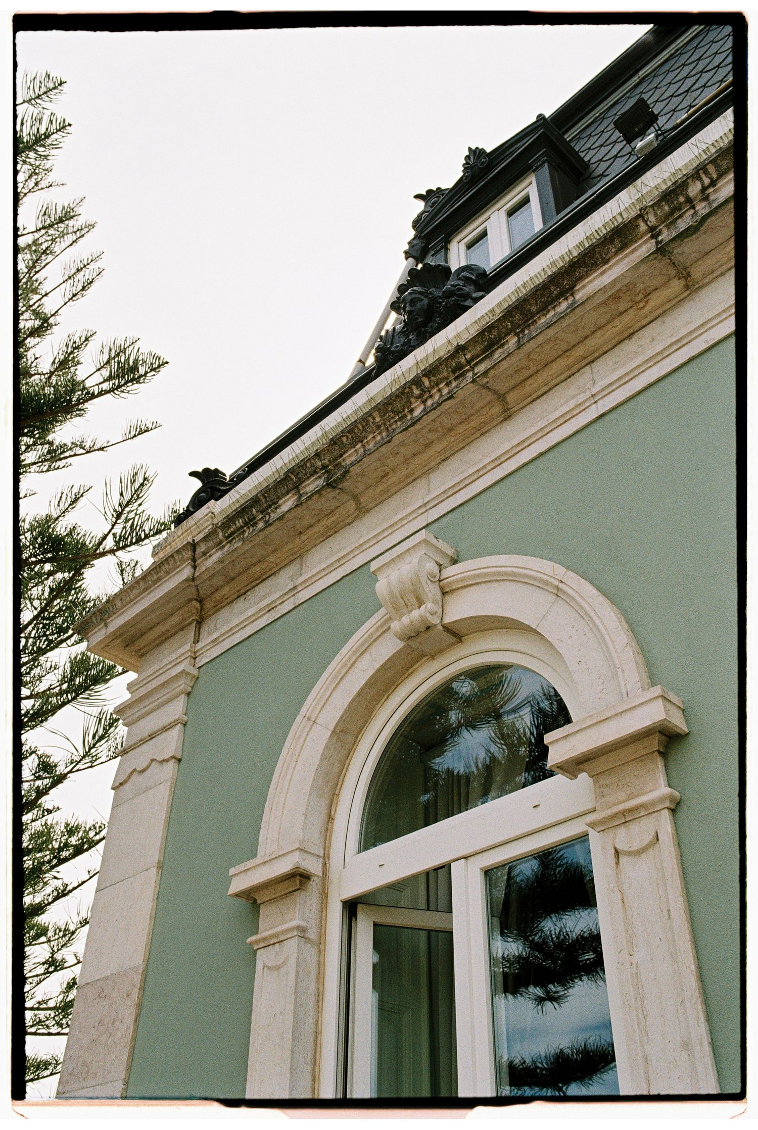 Historic European building with ornate arched window, cream stone moldings, and mint green facade beneath a mansard roof.
