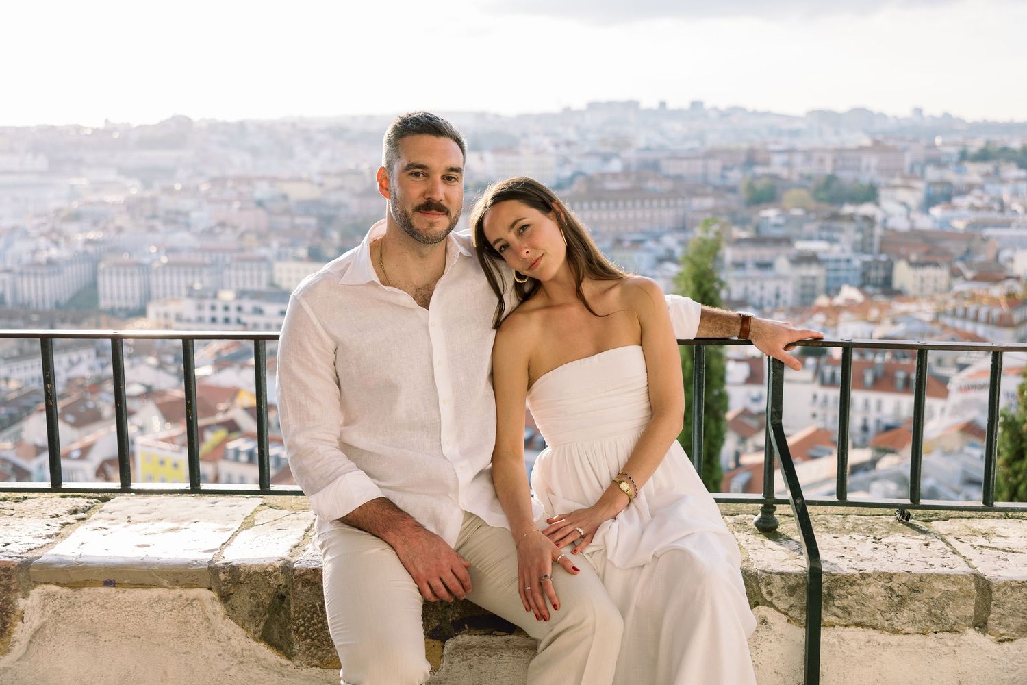 Couple in white outfits sitting on a stone wall with a panoramic cityscape view in the background.