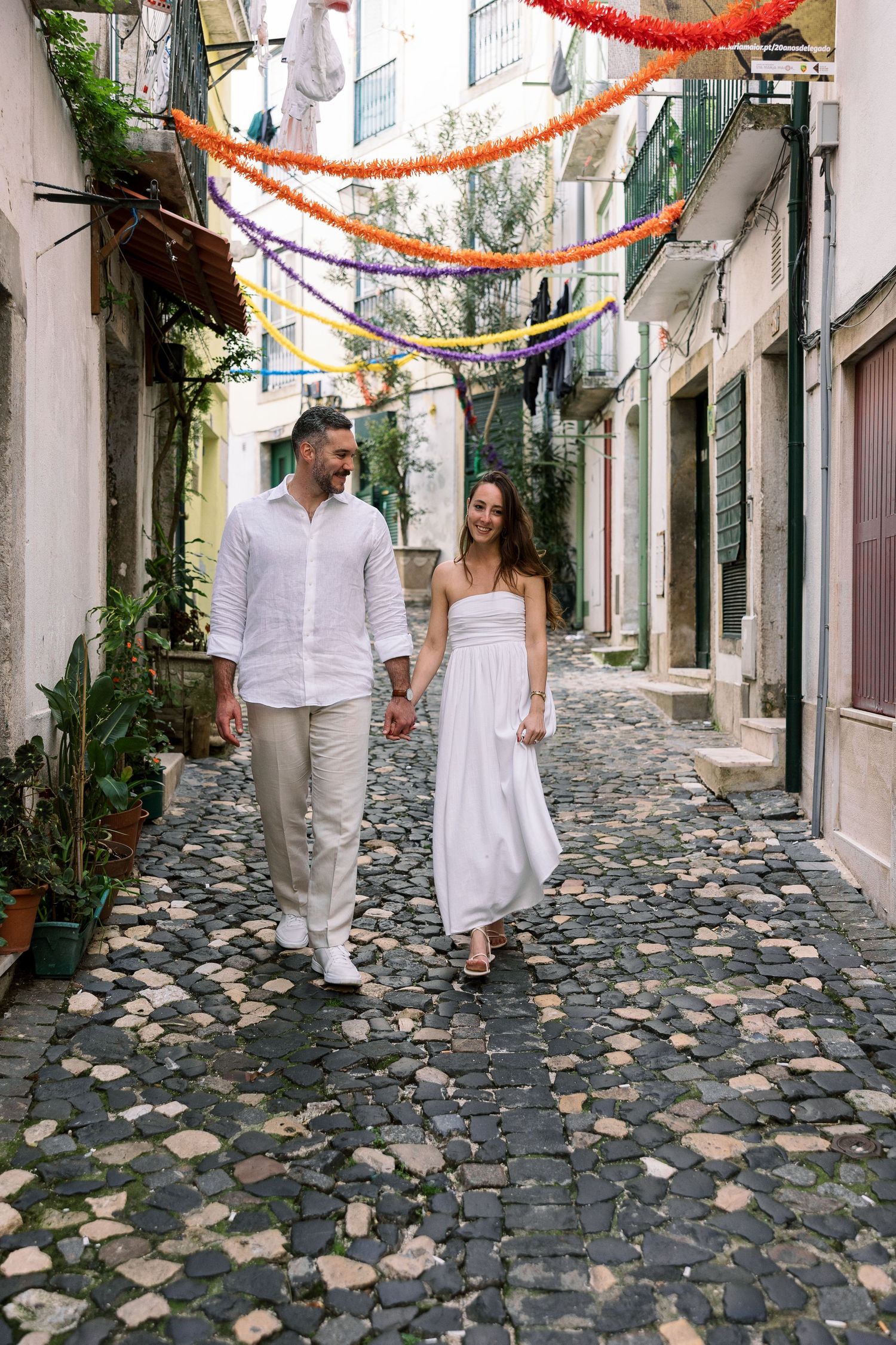 Couple in white outfits holding hands walking on cobblestone alley with colorful garlands in a European old town.