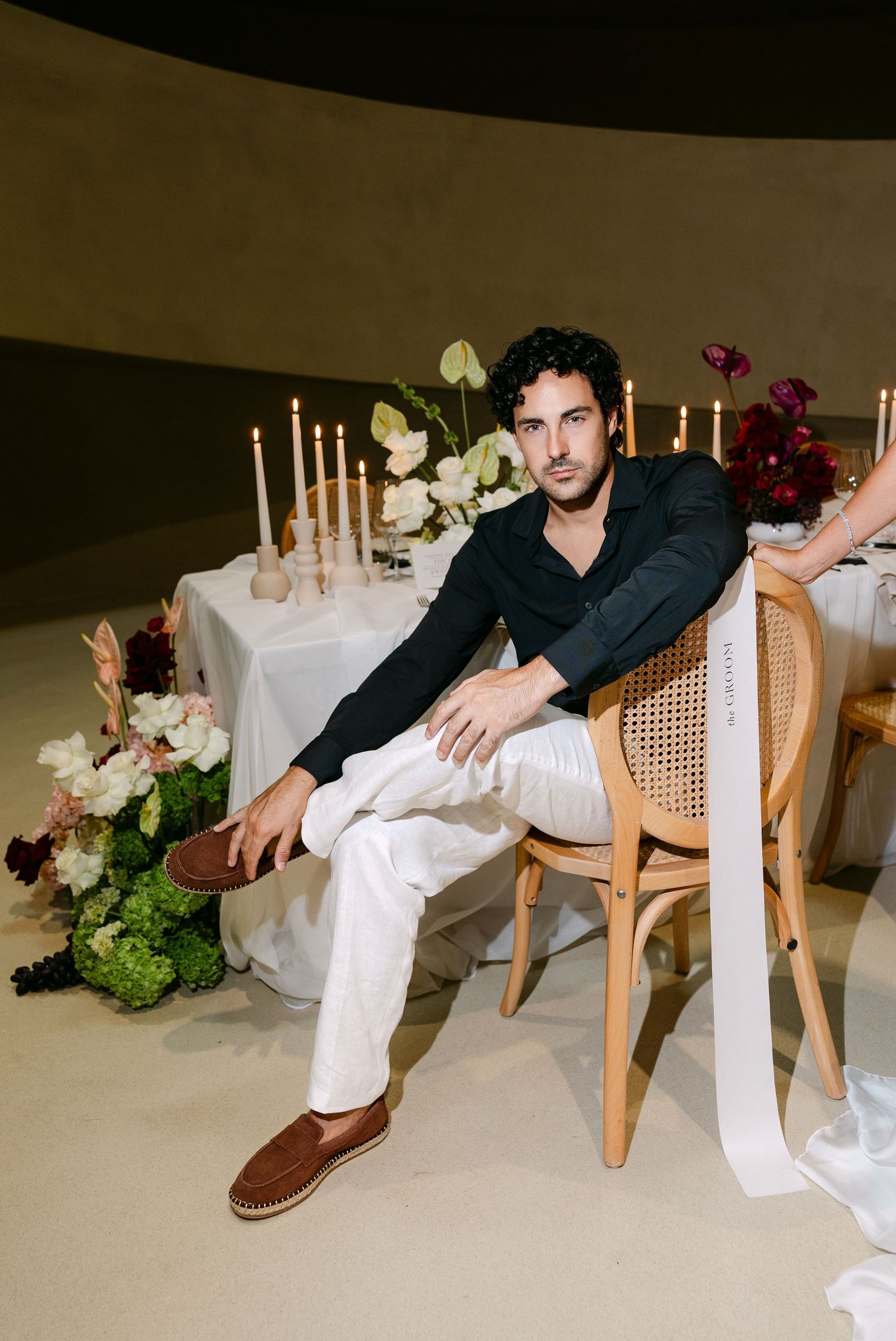 Groom in black shirt and white pants sits relaxed at candlelit wedding reception table with florals.