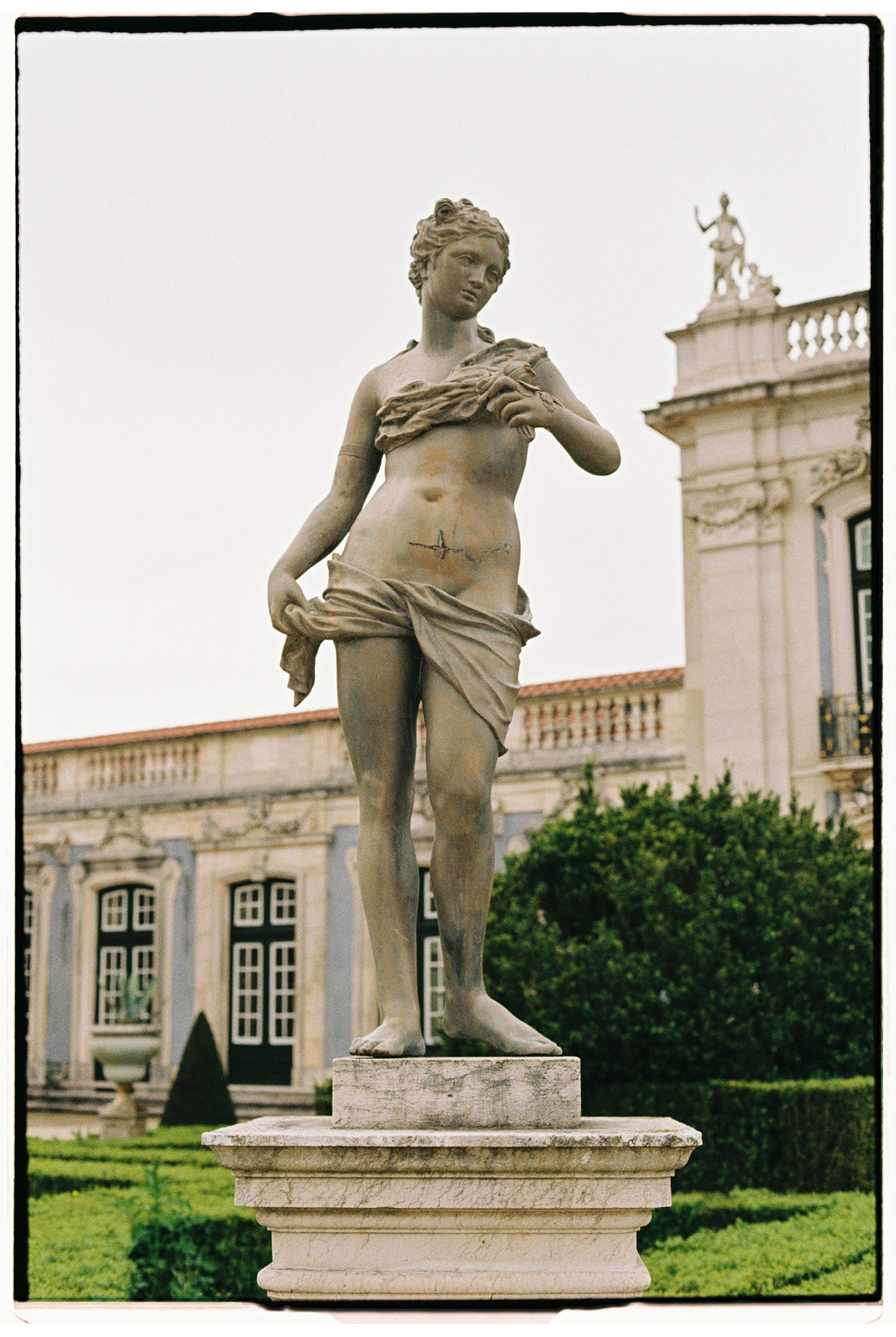 Classical stone statue of a semi-draped female figure on pedestal in formal garden with ornate building facade.