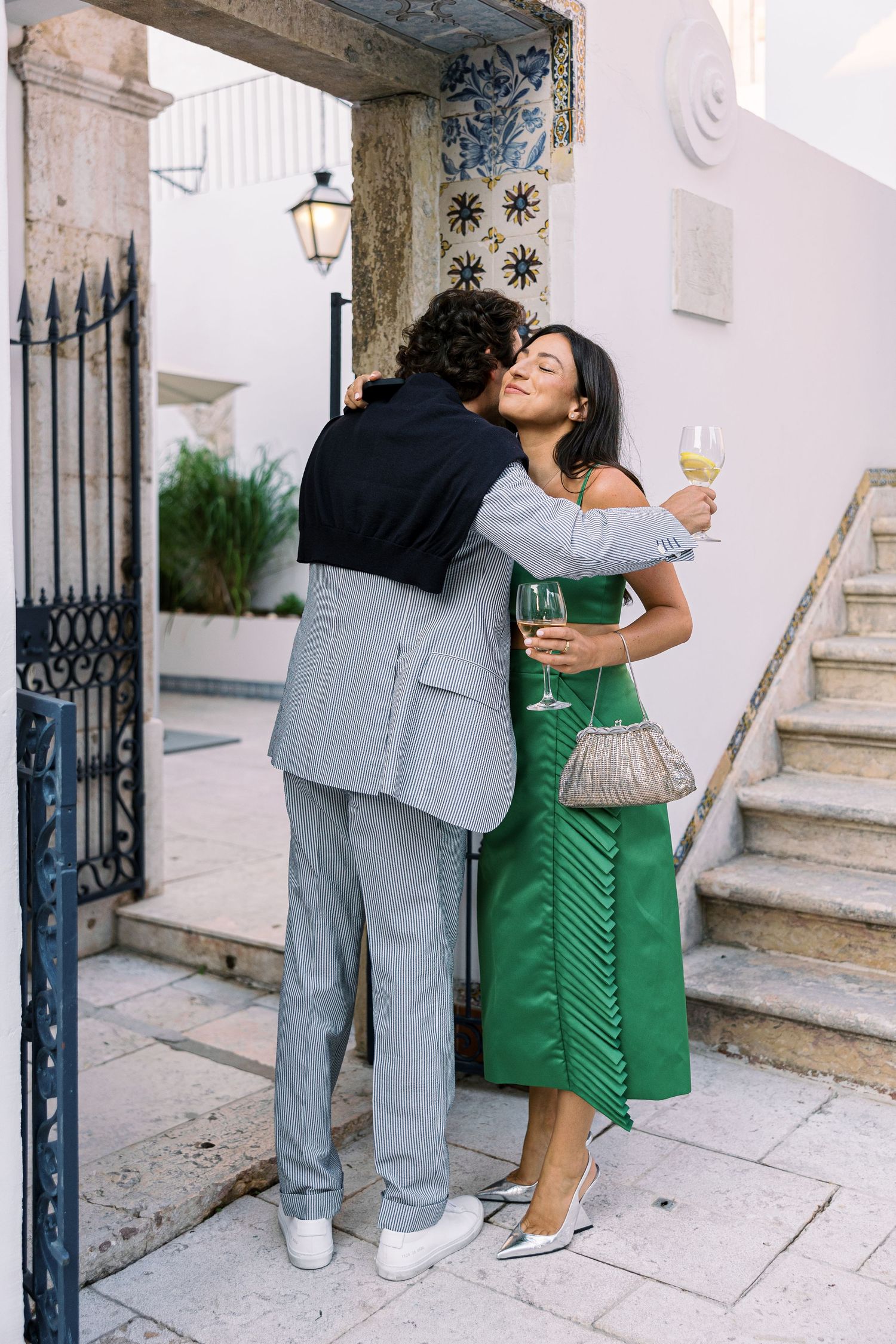 Couple embracing outdoors, woman in green dress holding champagne, man in gray suit, beside ornate gate and tiled wall.