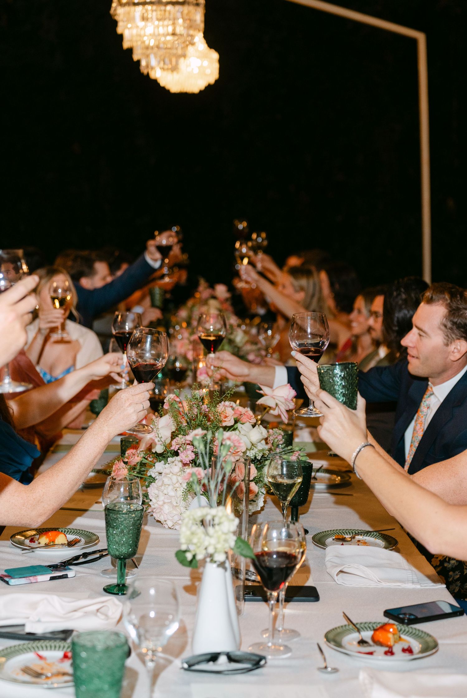 Wedding guests raise glasses in toast under crystal chandelier at outdoor reception.