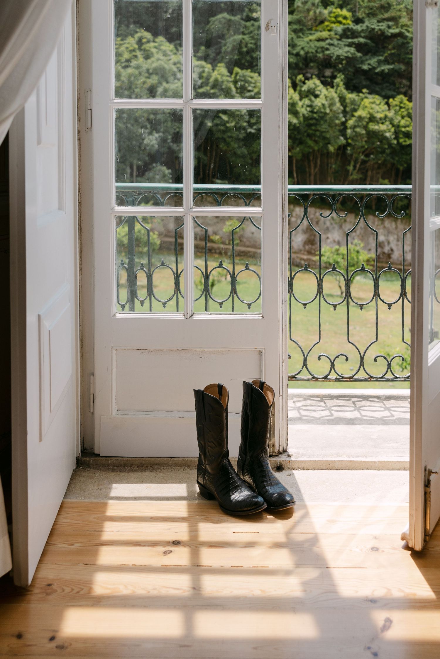 A pair of black boots sits in sunlit doorway with decorative iron railing visible through window.