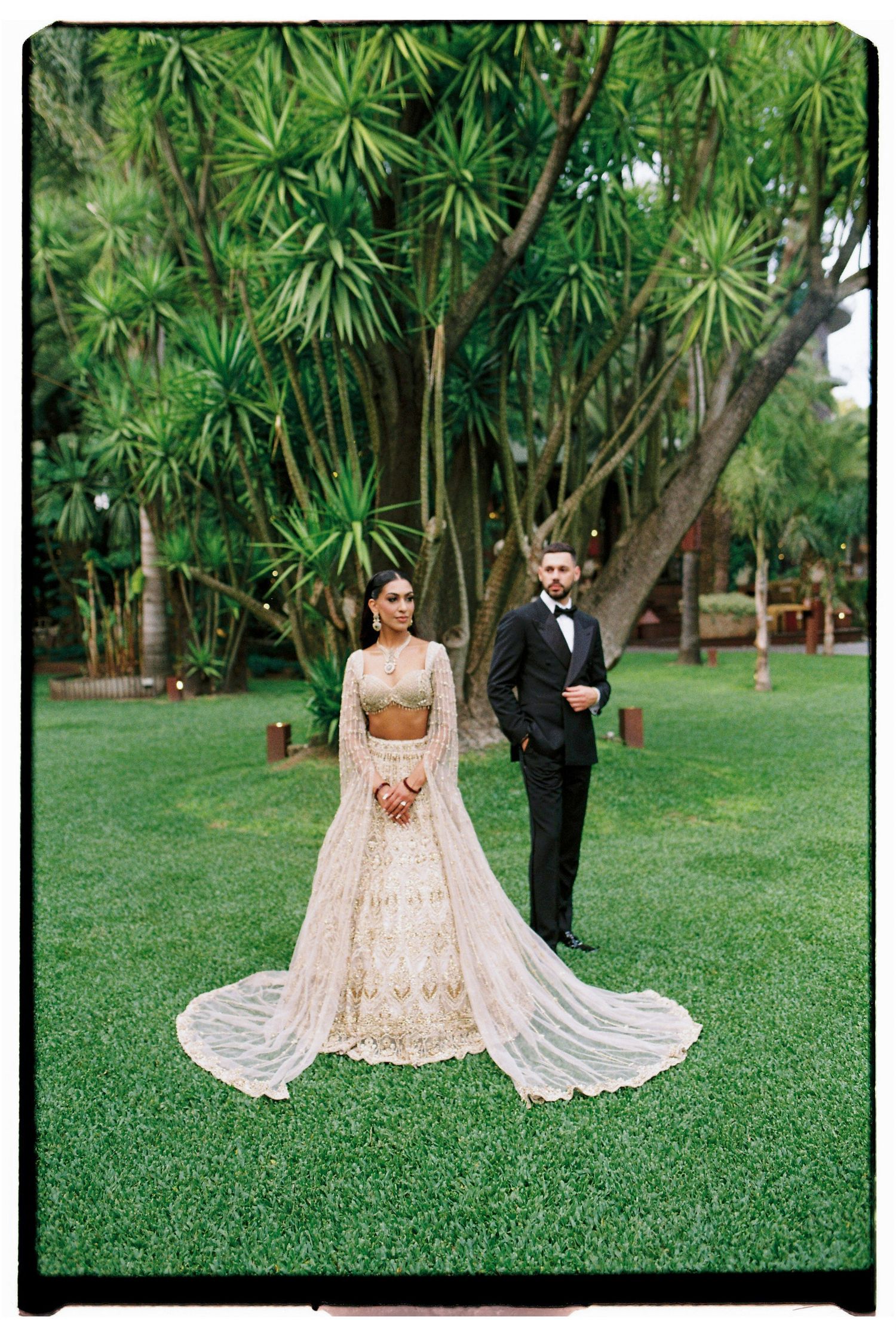 Bride in gold lehenga and groom in black tuxedo pose on lush green lawn surrounded by tropical trees.
