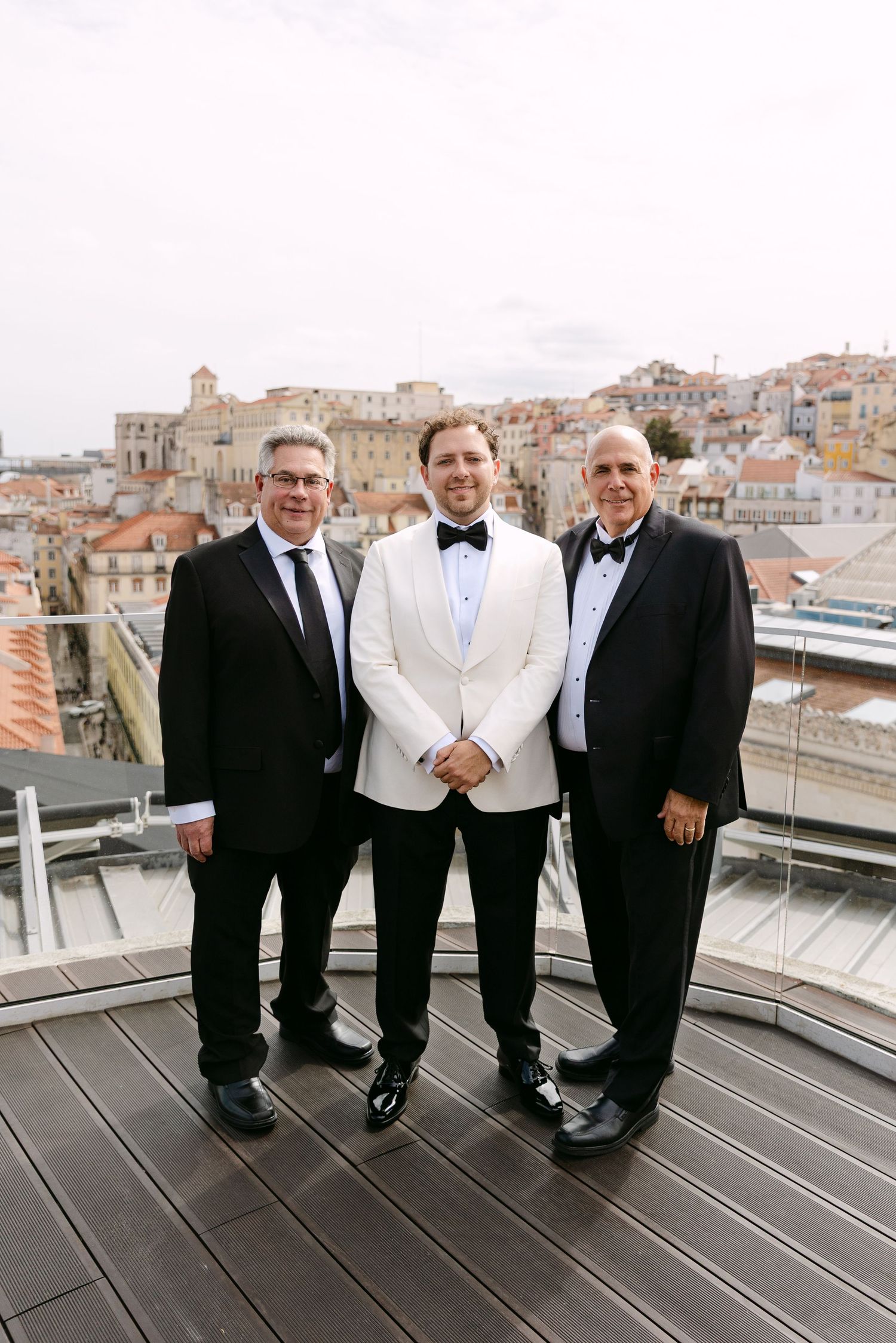 A group in formal attire poses on a rooftop terrace with city buildings in the background.