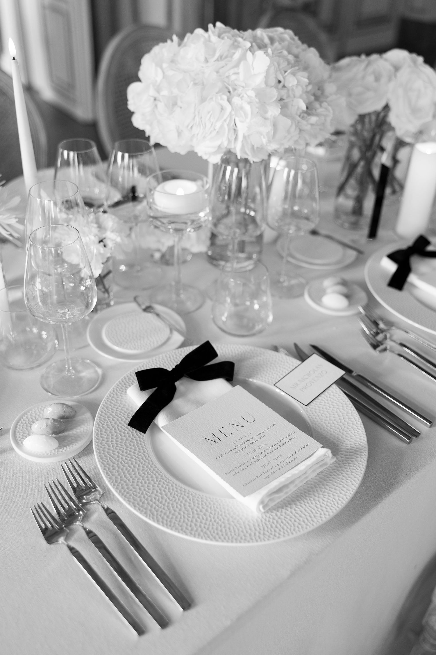 Elegant table setting with white flowers, crystal glasses, and menu card tied with black bow.
