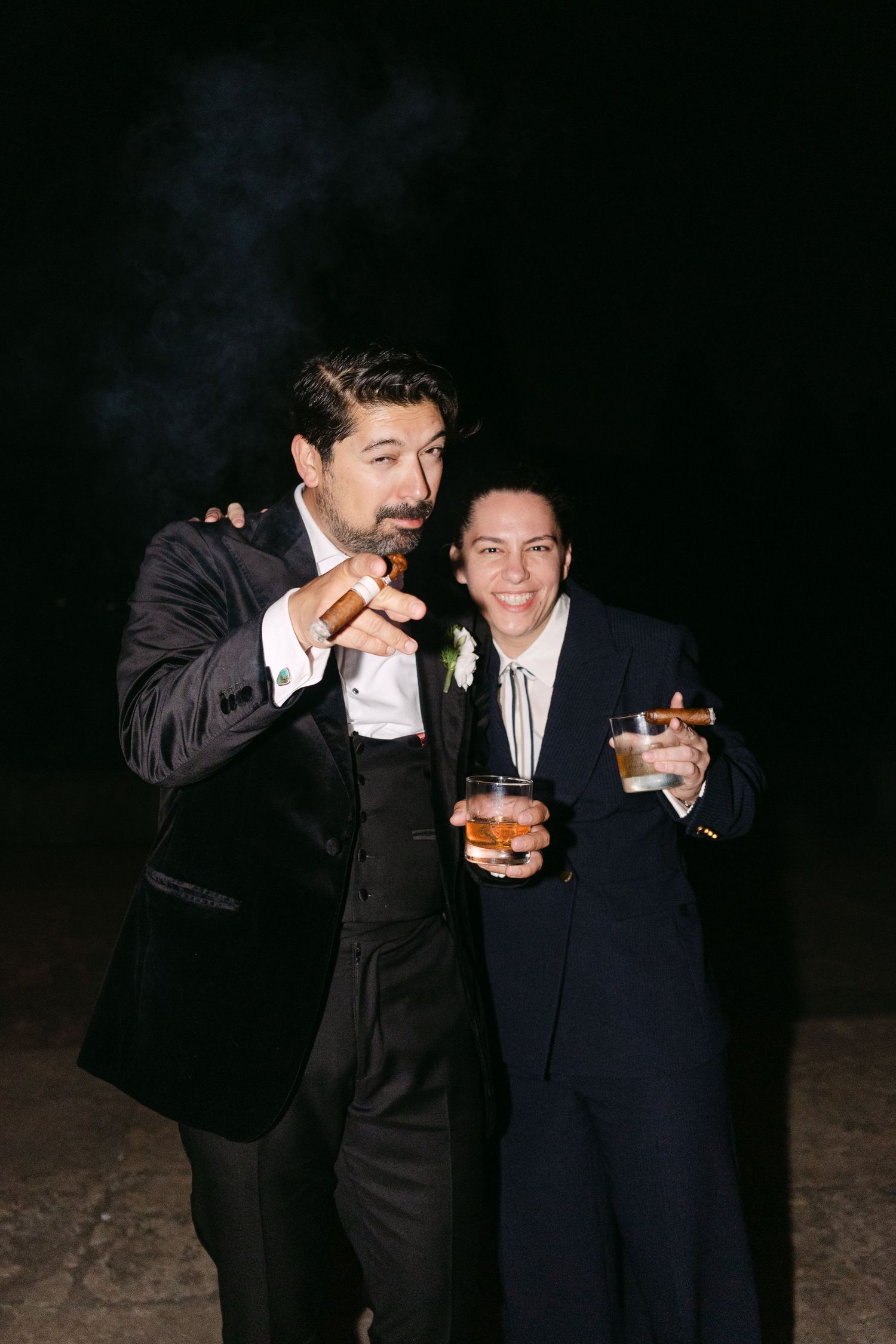 Two guests in tuxedos enjoying cigars and drinks at an evening wedding reception.