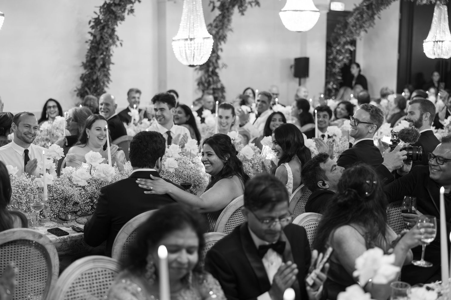 Black and white photo of wedding guests enjoying an elegant reception dinner under chandeliers.
