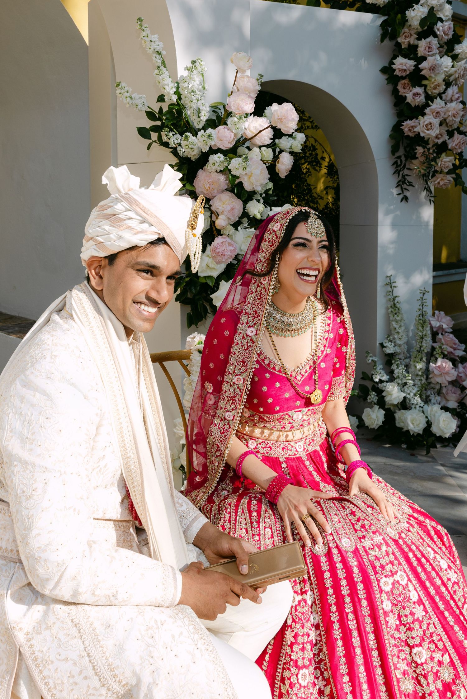 Newlyweds in traditional Indian wedding attire share joyful moments under white floral arch.