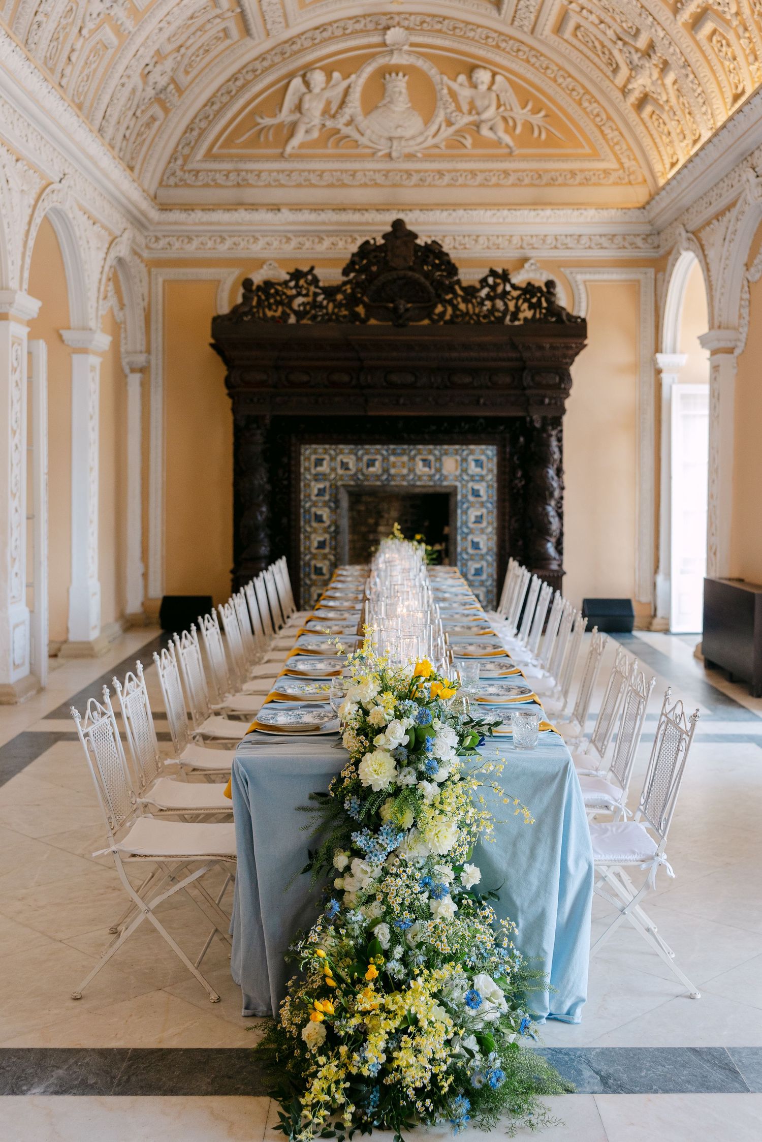 Formal dining table with blue tablecloth and floral arrangements in an ornate white classical room.