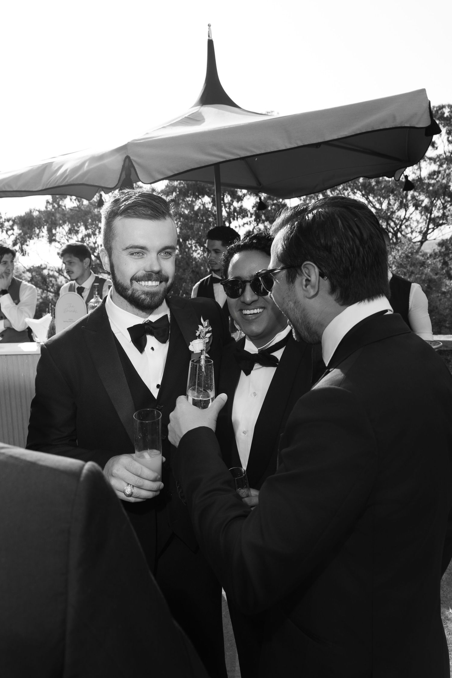 Groomsmen in black tuxedos socialize under outdoor tent at wedding reception.
