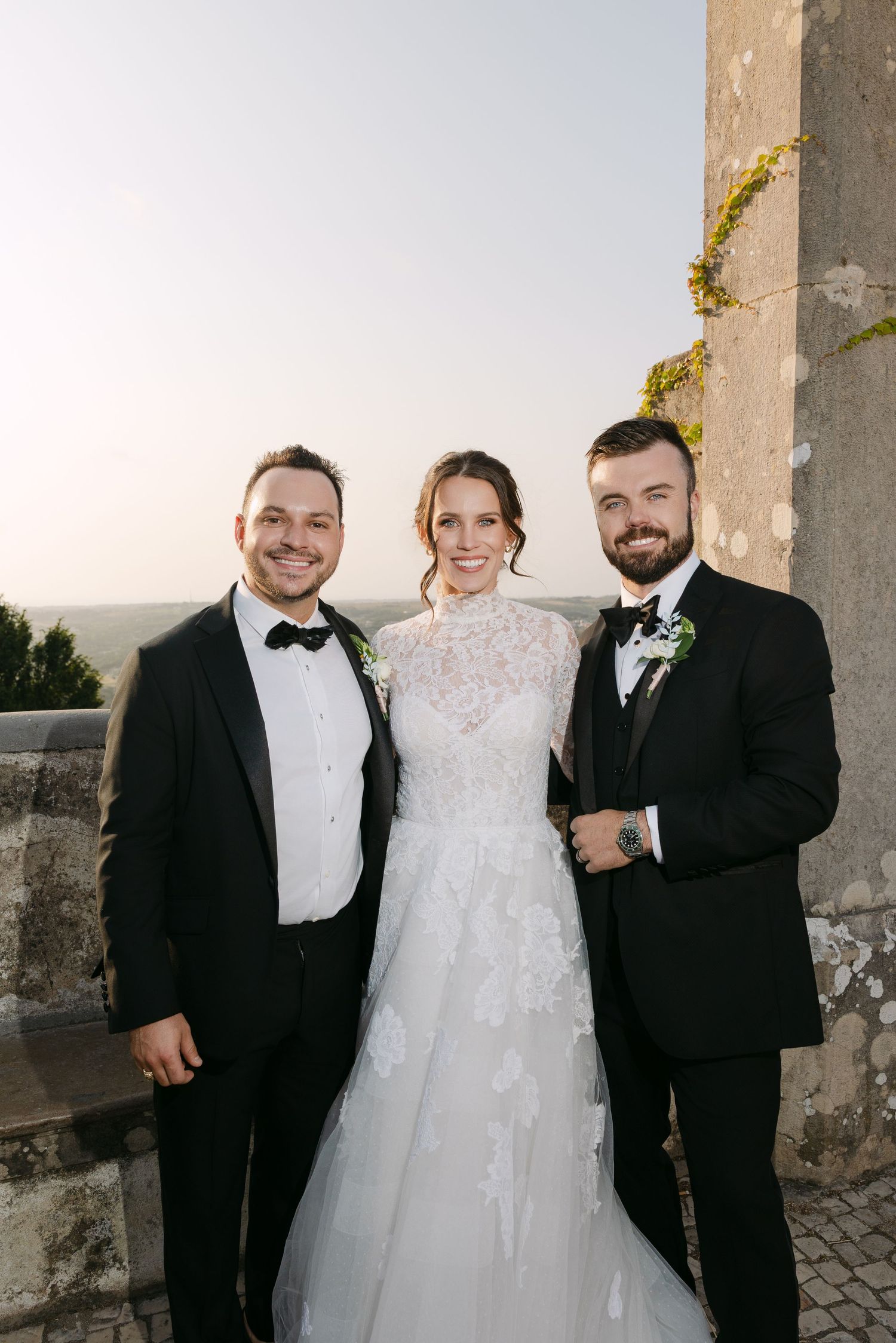 Wedding party poses together in formal attire against stone architectural backdrop.