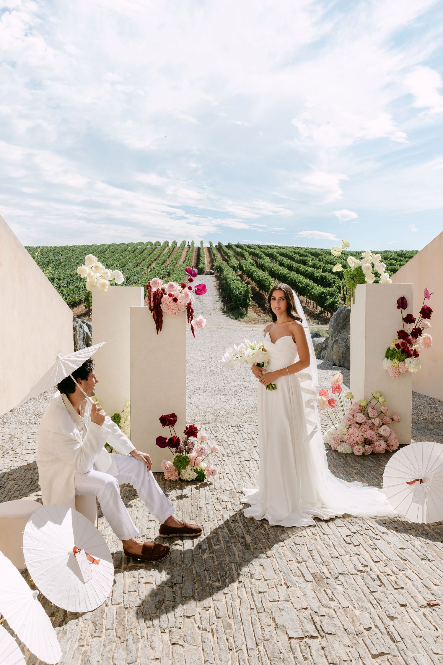 Bride and groom pose among white panels adorned with colorful flowers at an outdoor vineyard wedding ceremony.