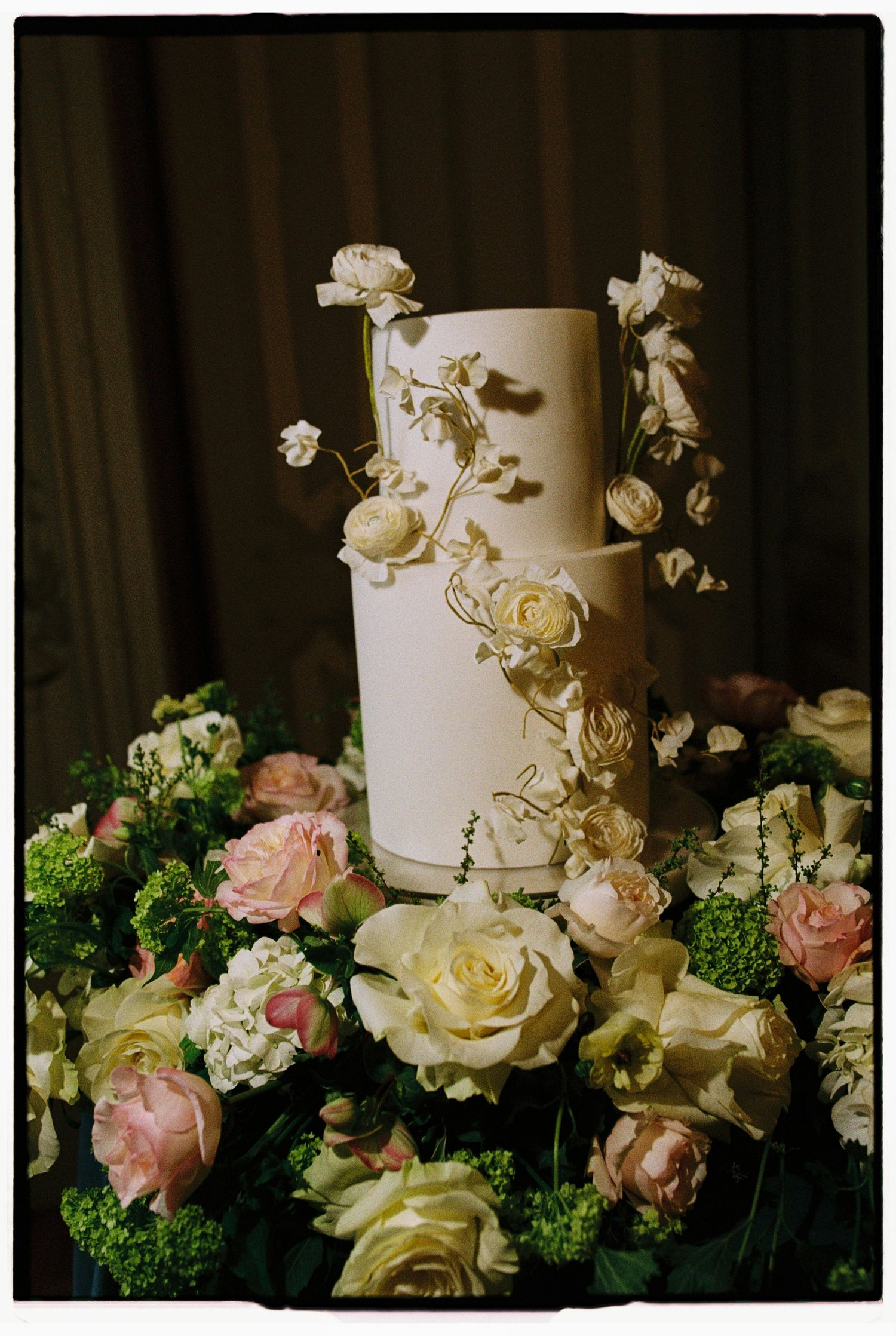Elegant white wedding cake adorned with gold floral details, surrounded by pink and cream roses and lush greenery.