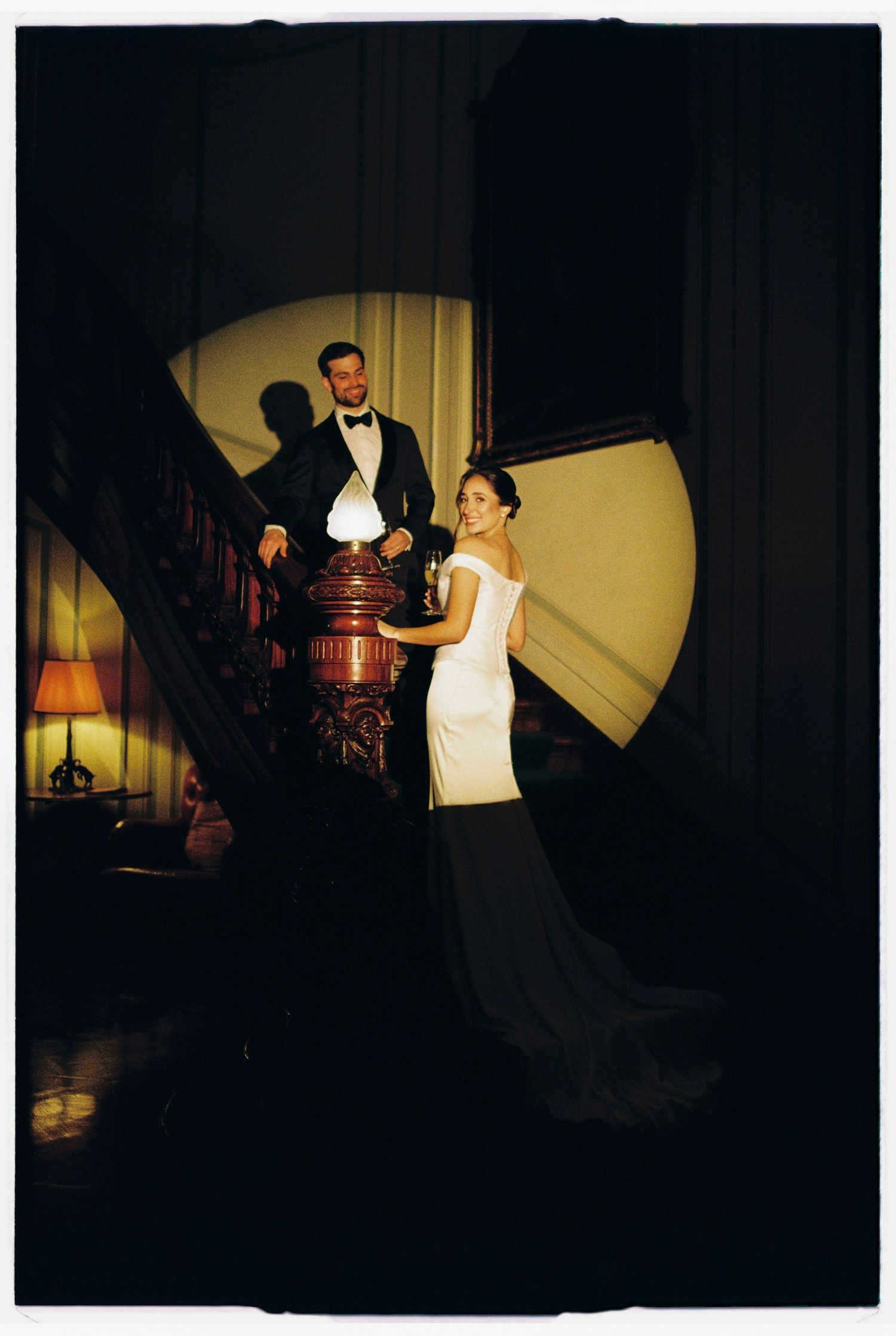 Elegant bride in white gown and groom in tuxedo posing on grand staircase in dimly lit venue.