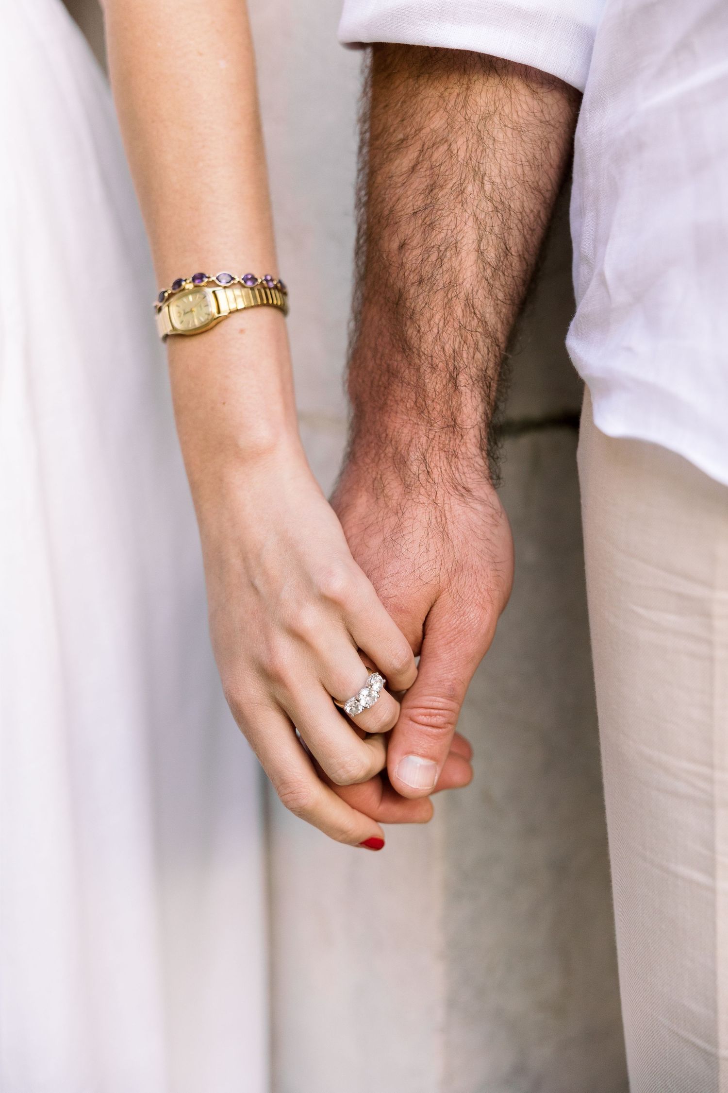 Close-up of a couple holding hands, woman wearing a diamond engagement ring and gold watch, man in white shirt.