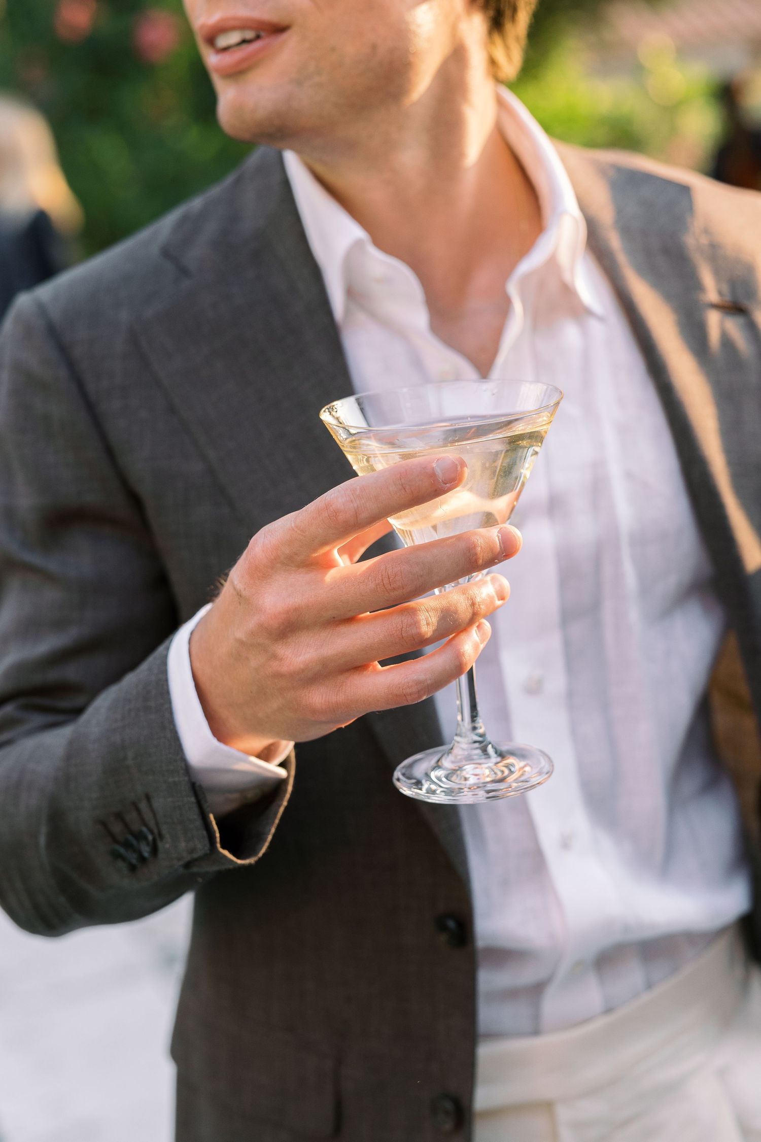 Man in gray blazer holding crystal martini glass at outdoor event.