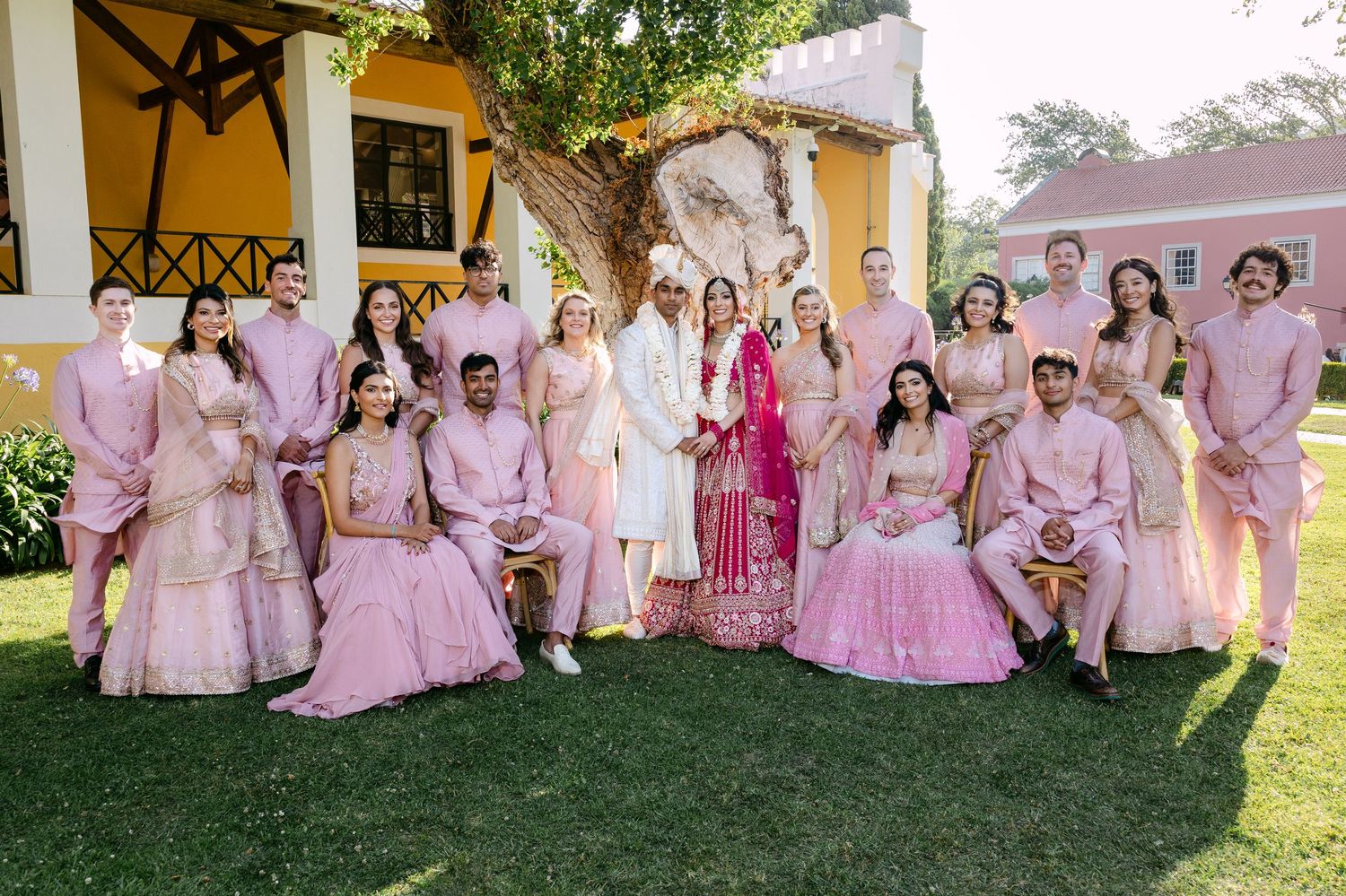 Wedding party in pink attire poses together on a grassy lawn during sunset.