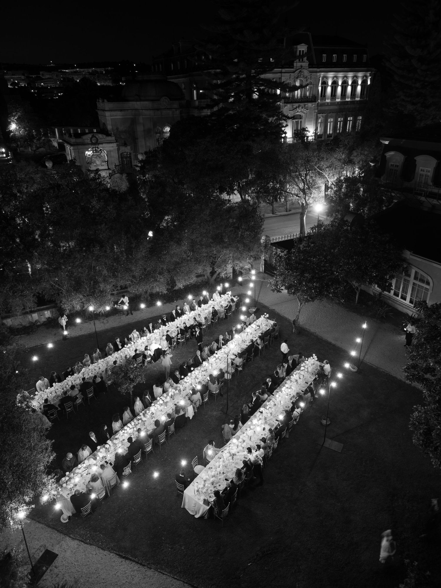 Aerial view of an outdoor wedding reception with long tables illuminated by string lights at night.