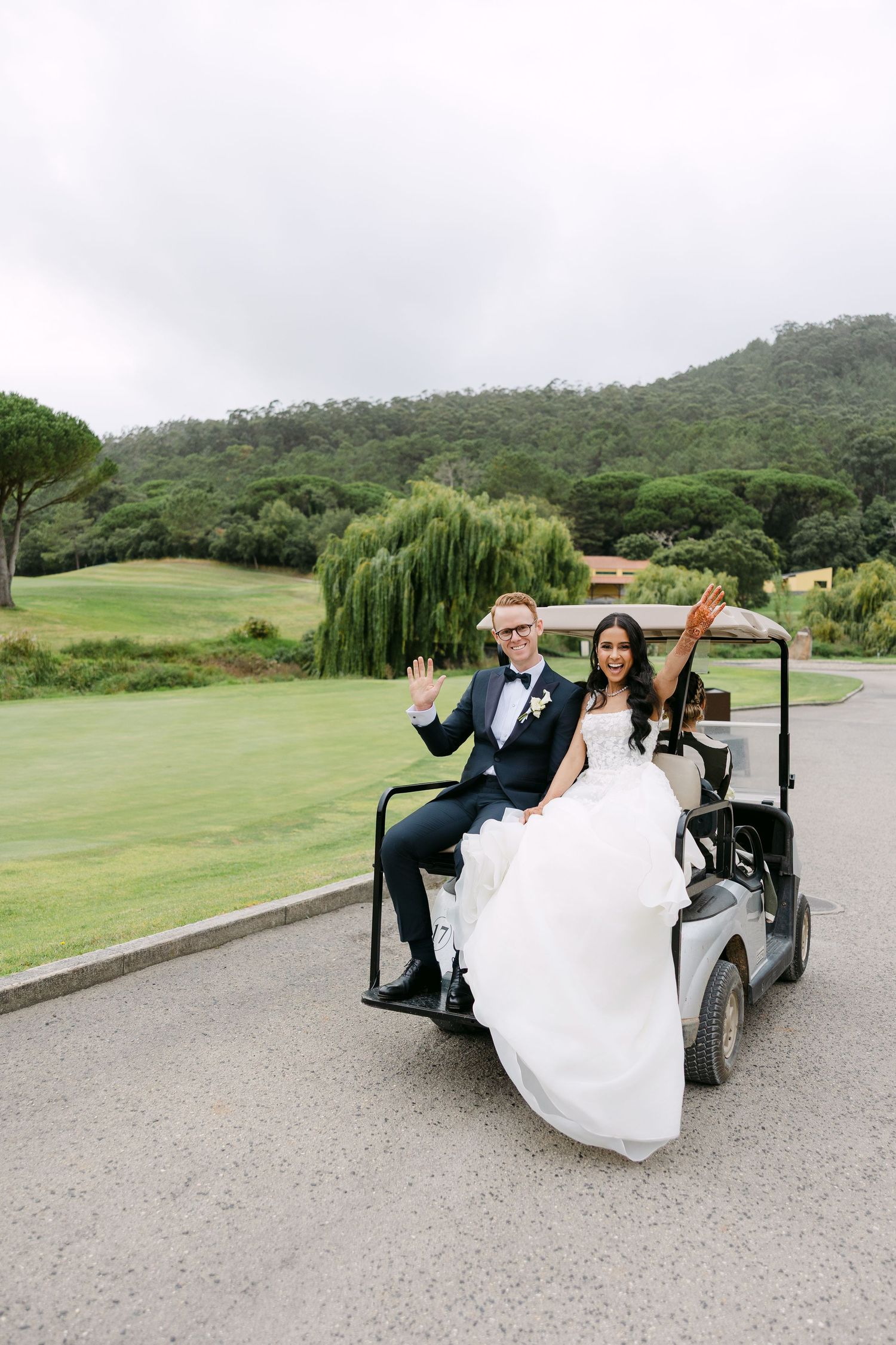 Newlyweds ride together in a golf cart on their wedding day across lush green grounds.