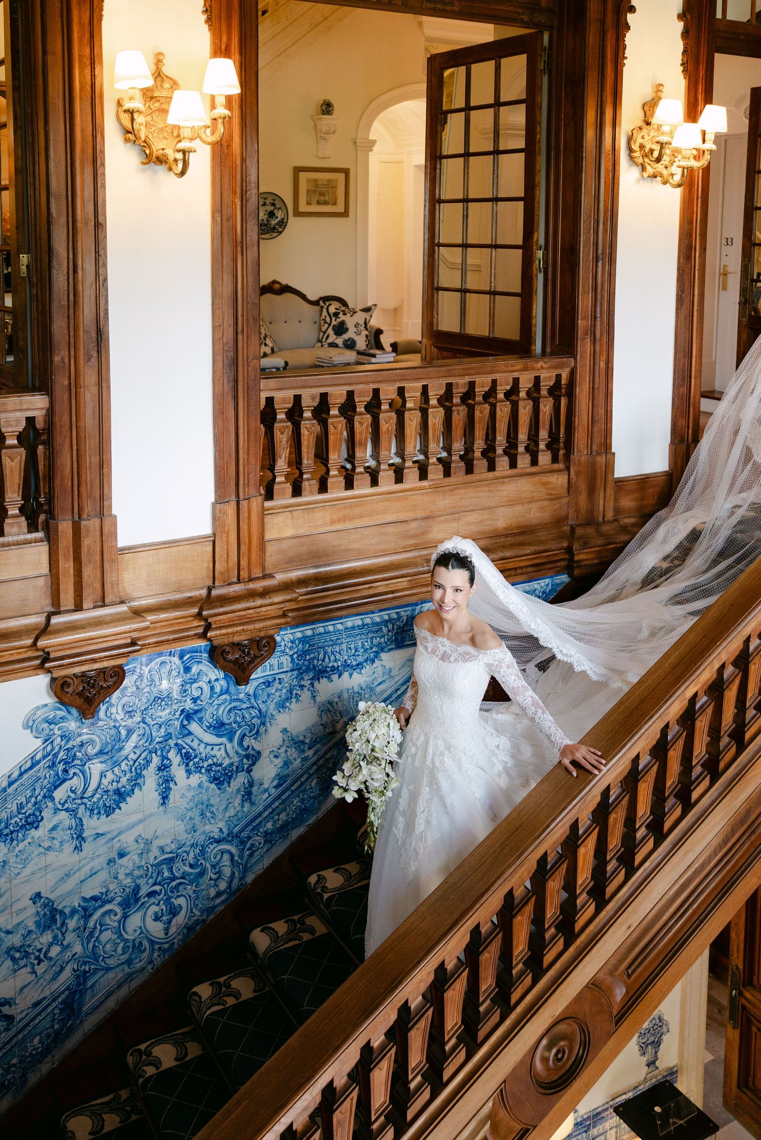 An elegant wedding dress descending a grand staircase with ornate blue tile walls and wooden railings.