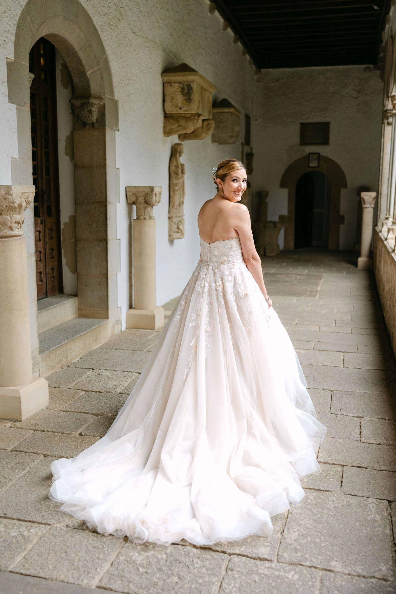 Flowing white wedding gown sweeps across stone floors in historic monastery hallway.
