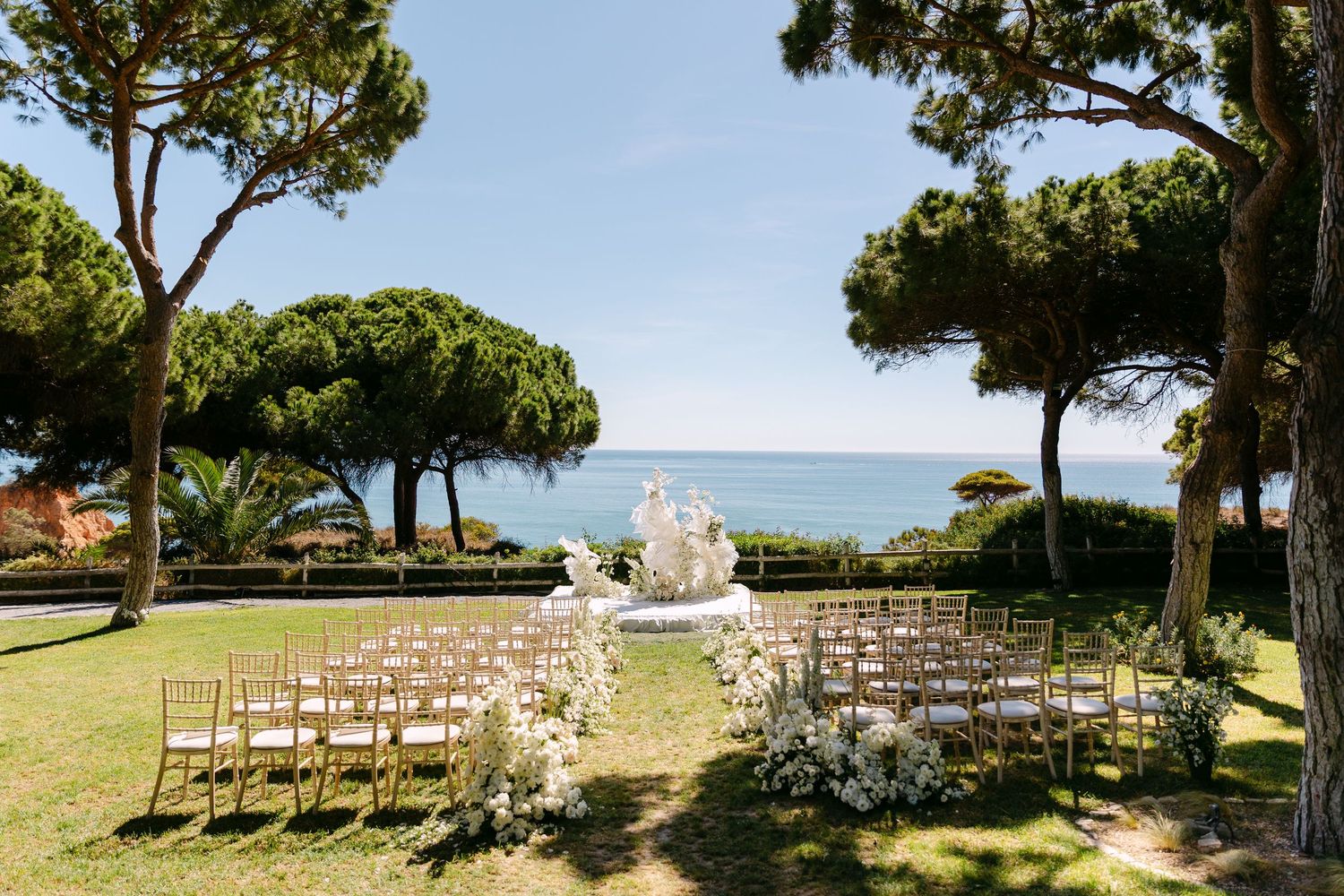 Outdoor wedding ceremony setup with wooden chairs facing the ocean under tall pine trees.