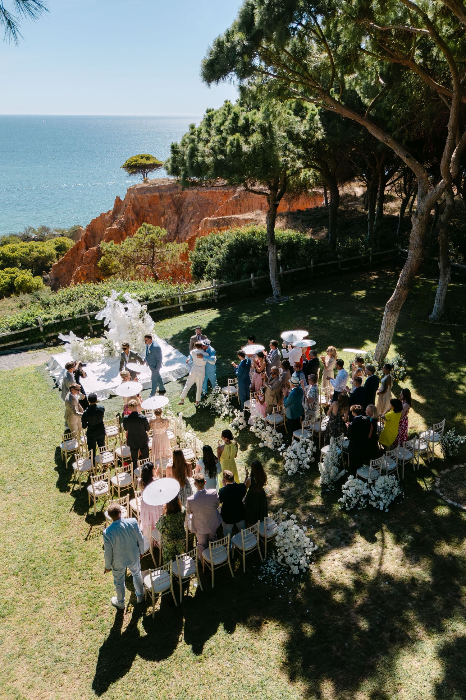Aerial view of an outdoor wedding ceremony by the ocean with guests gathered under trees.