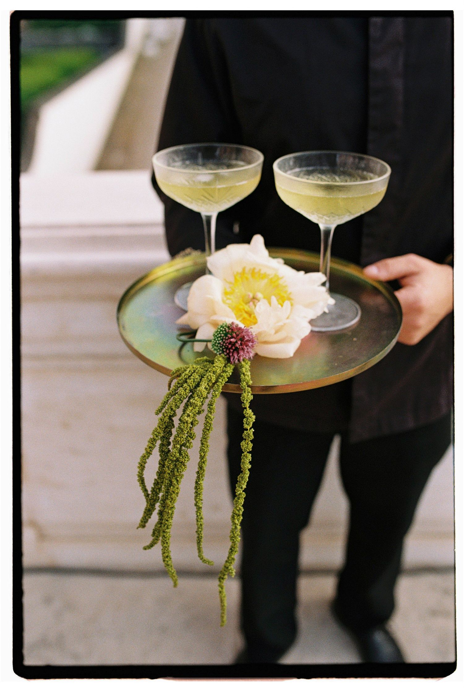 Waiter in black holding a gold tray with two coupe cocktails and a white peony flower arrangement.