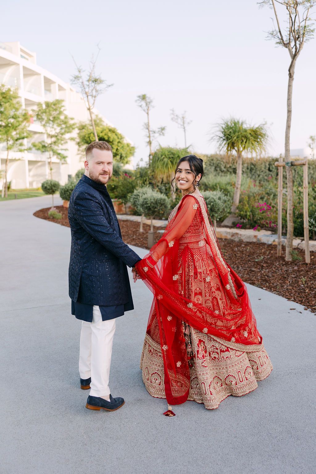 A couple poses outdoors in formal attire with the bride wearing a stunning red and gold embroidered lehenga.