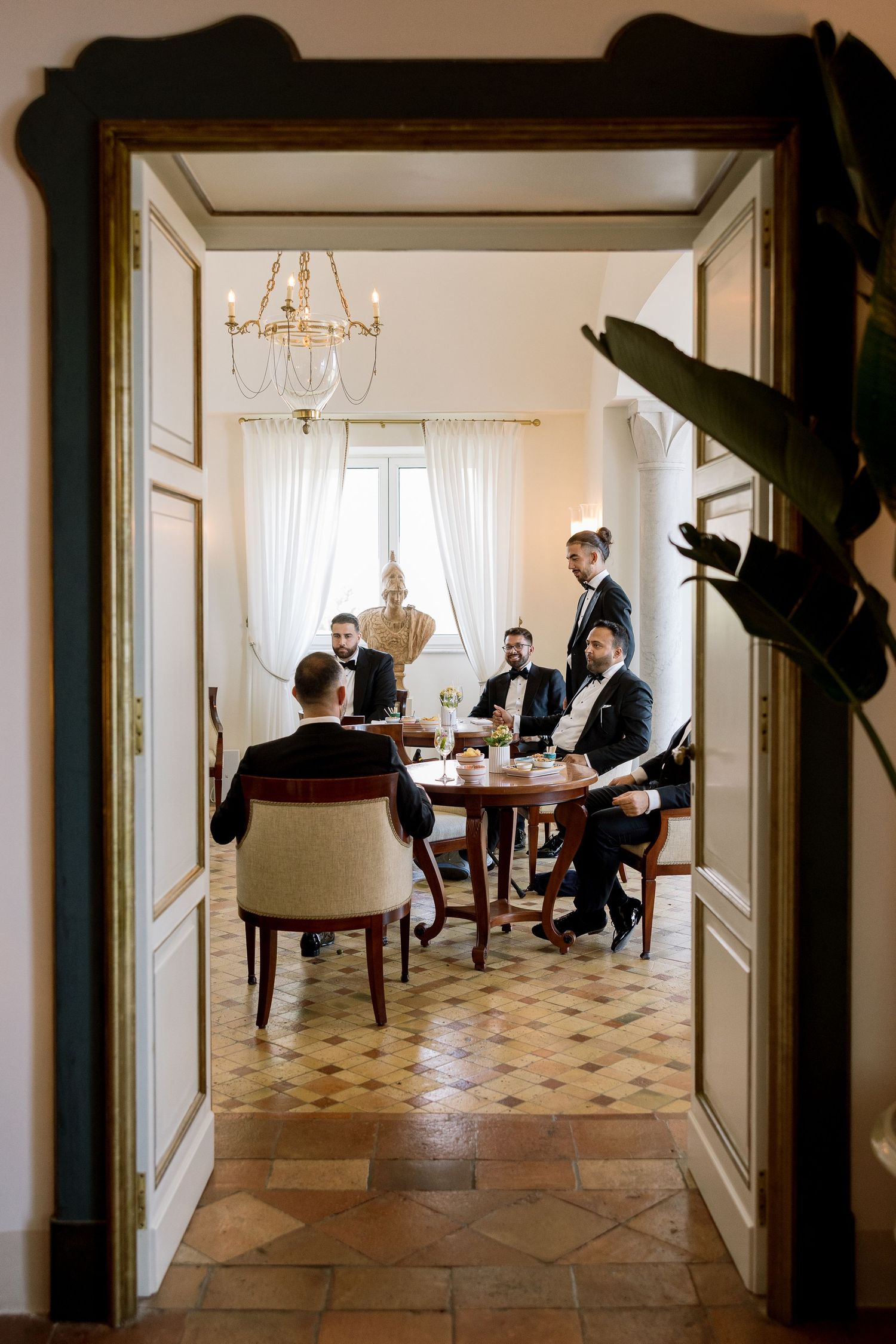 Elegant men in formal attire gathered around a antique table in a grand room with chandelier and ornate doors.