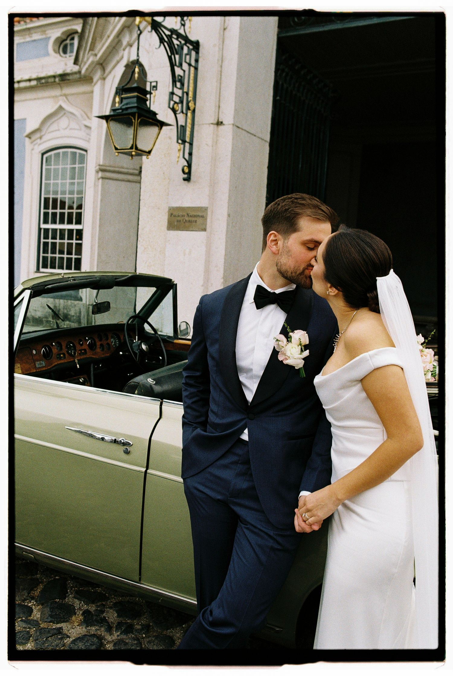 Bride and groom share a kiss leaning against a vintage green convertible car outside an elegant building.