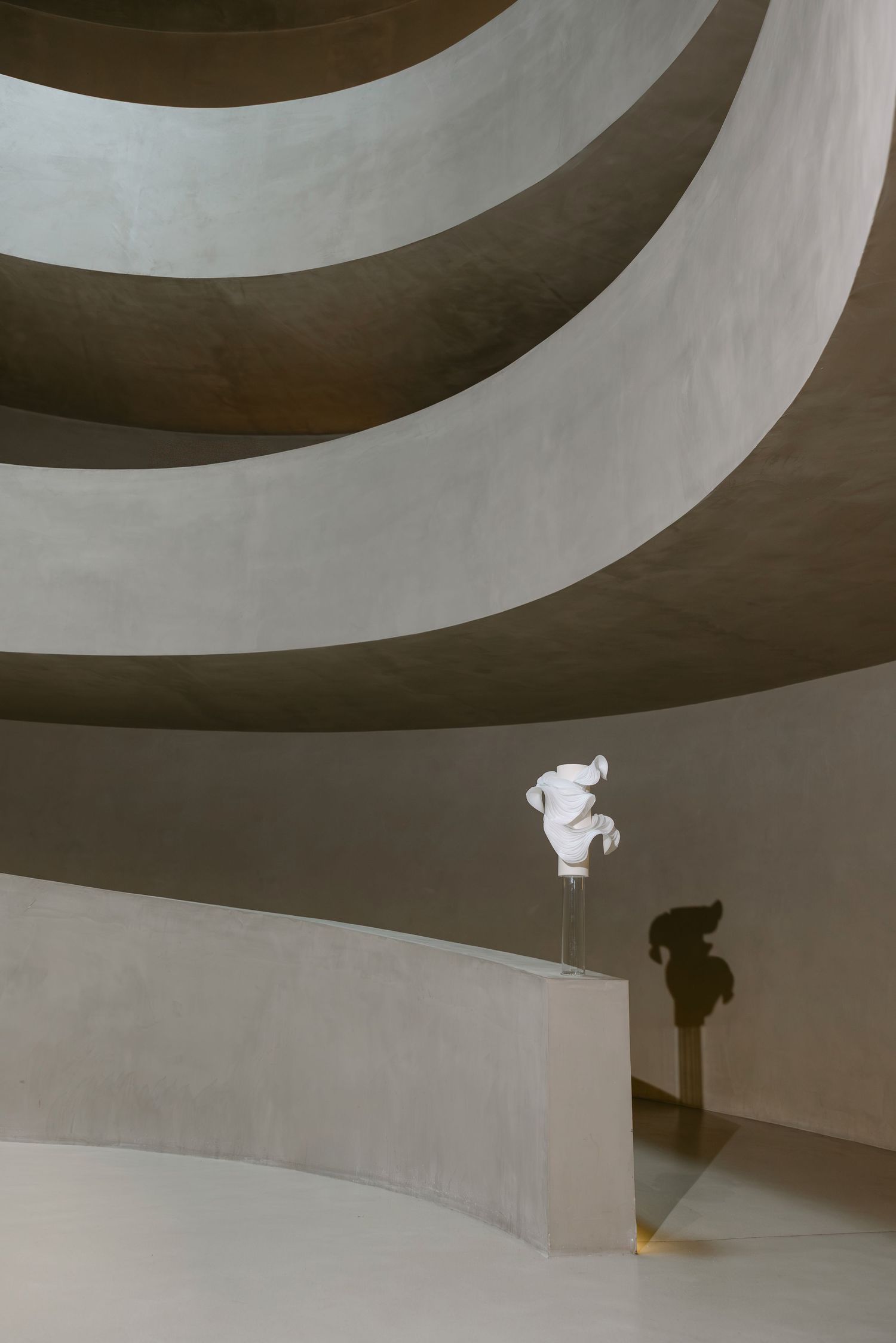 White abstract sculpture on pedestal in spiral concrete gallery with dramatic shadow on curved wall.