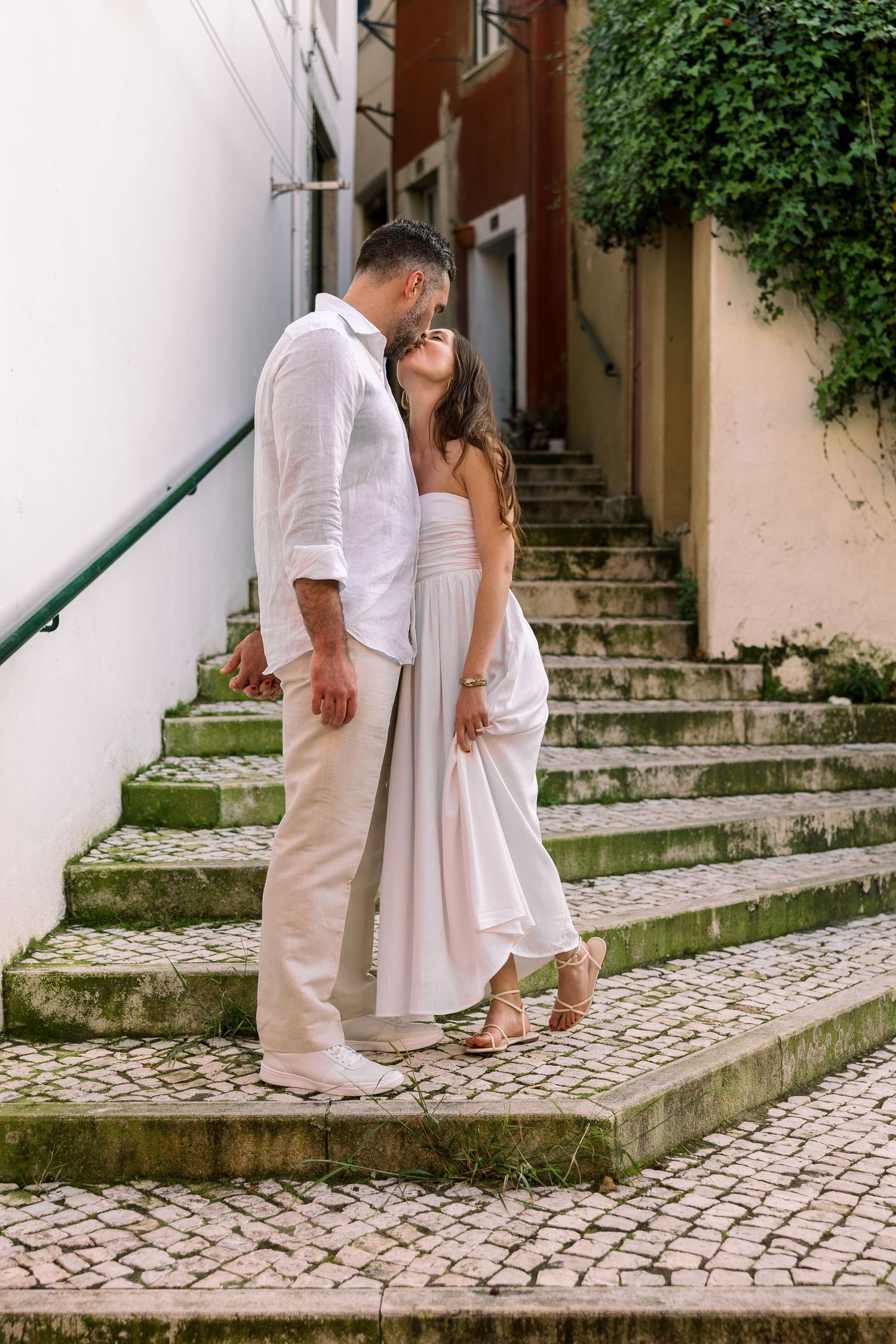 Romantic couple in white outfits sharing a tender moment on moss-covered stone steps in a charming European alley.