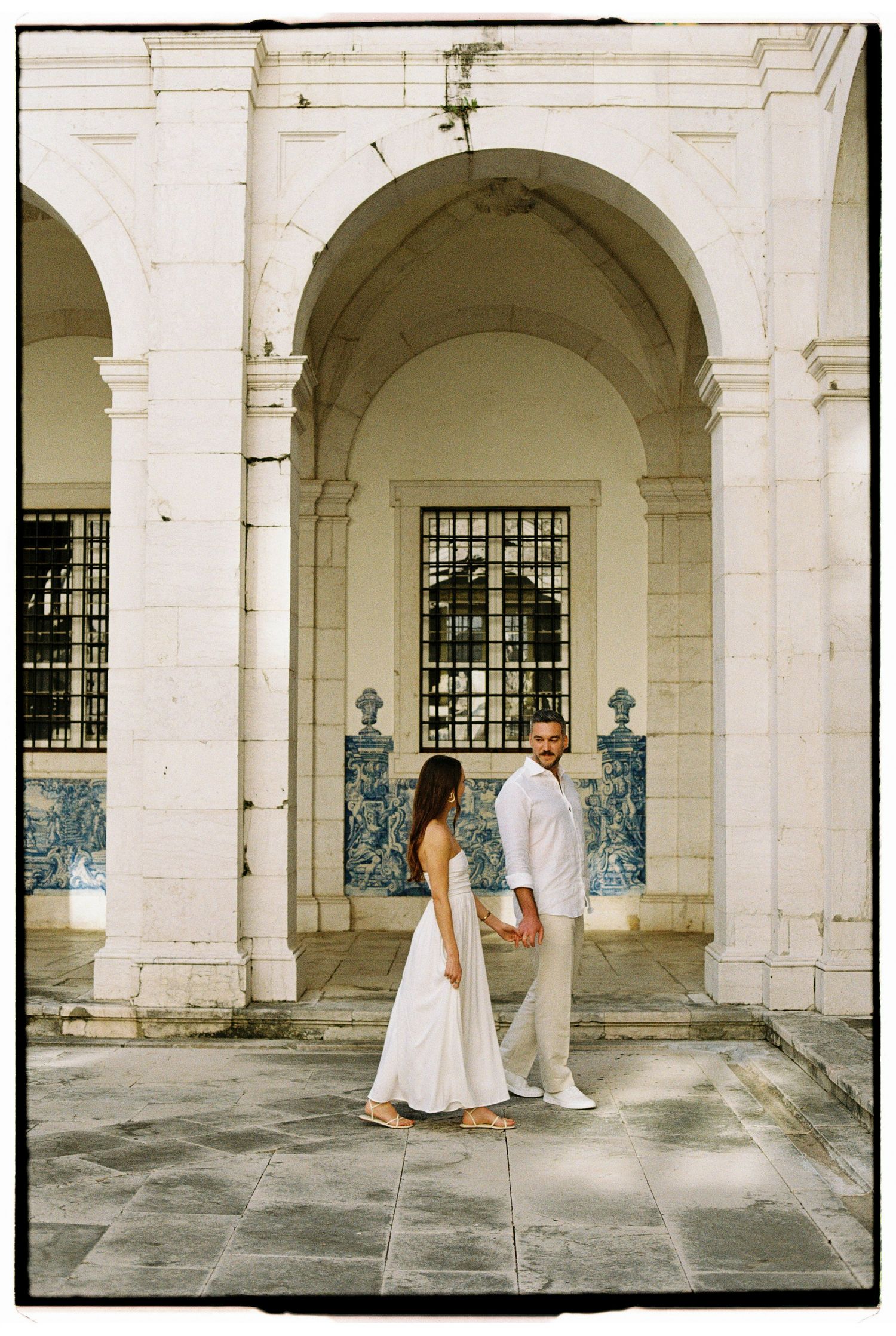Couple in white outfits holding hands before ornate stone archway with blue azulejo tiles.