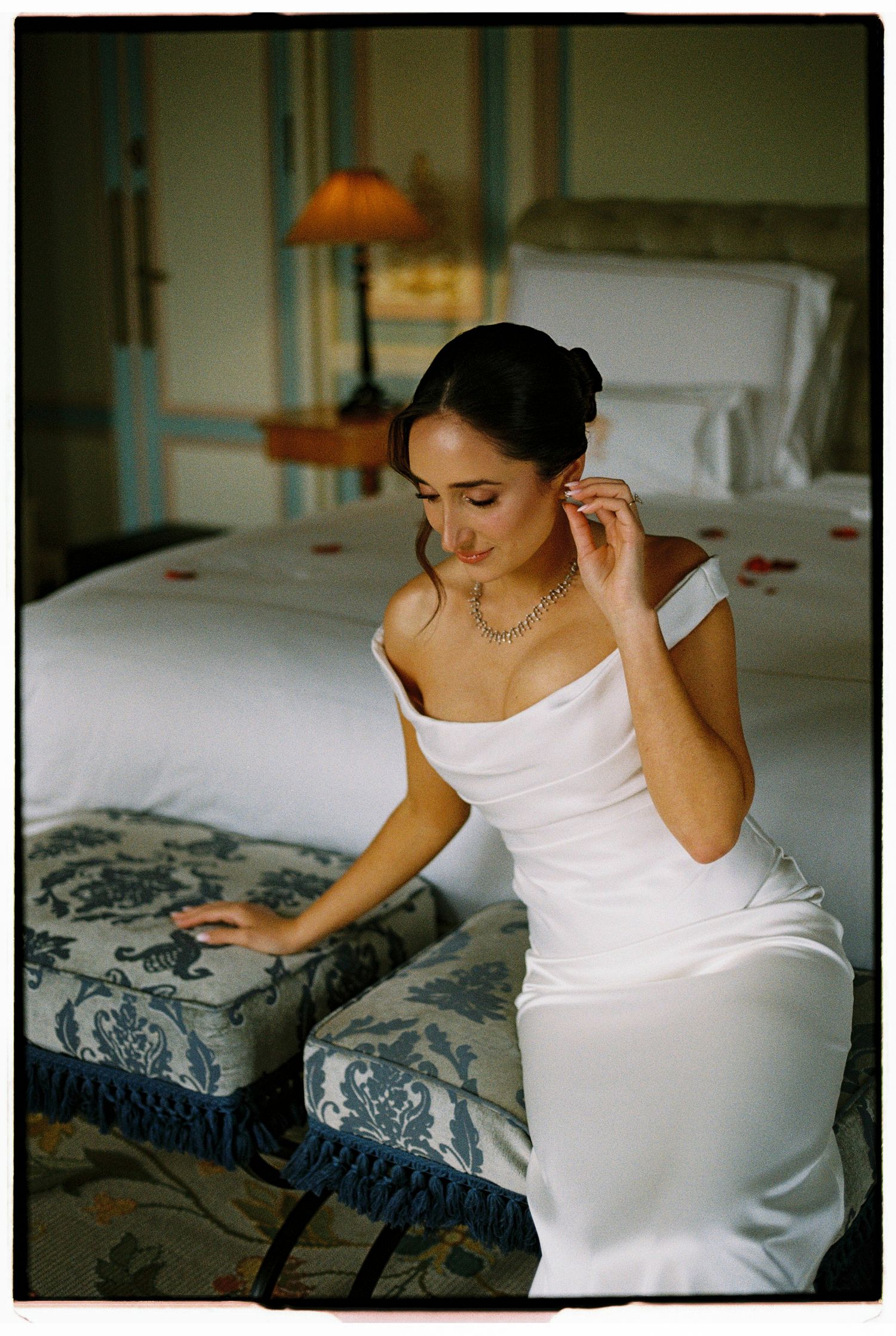 Elegant bride in white off-shoulder gown adjusting earring while seated in a luxurious hotel room.