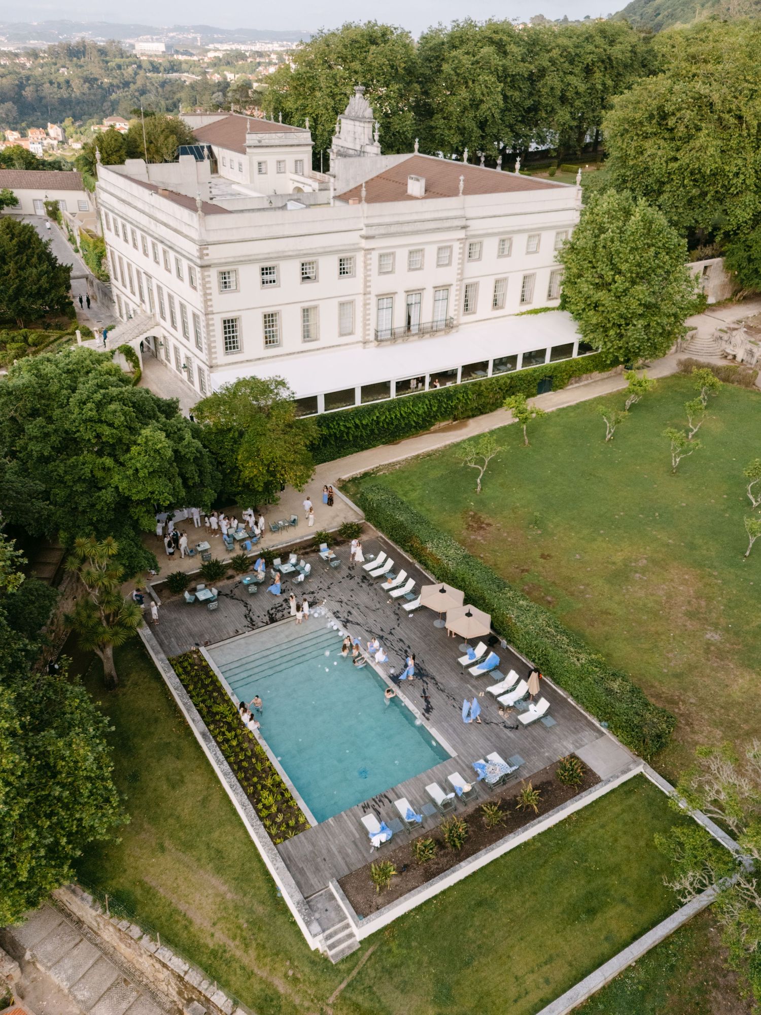 Aerial view of a grand white estate building with rectangular swimming pool and manicured grounds.