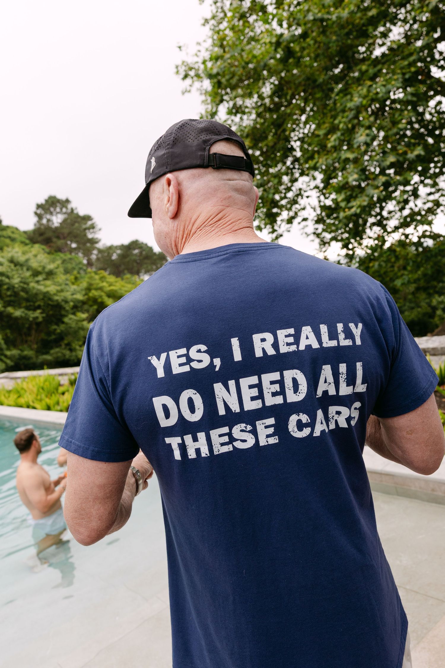 Man wearing navy t-shirt with humorous text about car collecting by a pool.