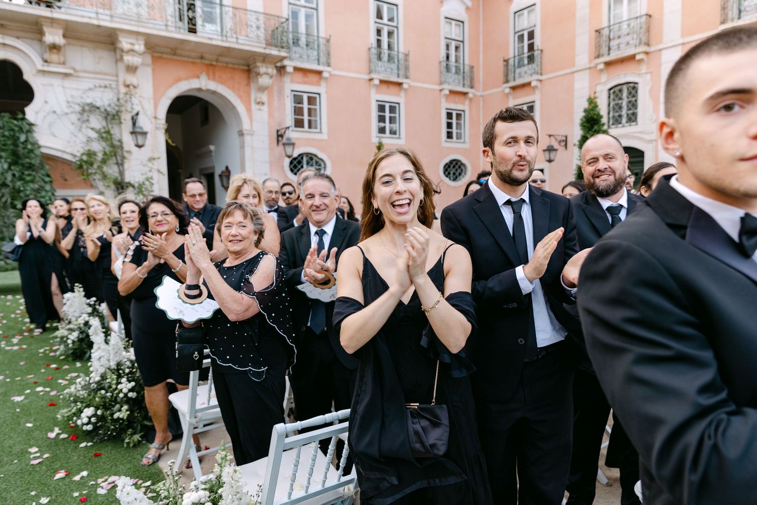 Wedding guests in formal attire applaud and celebrate during an outdoor ceremony.