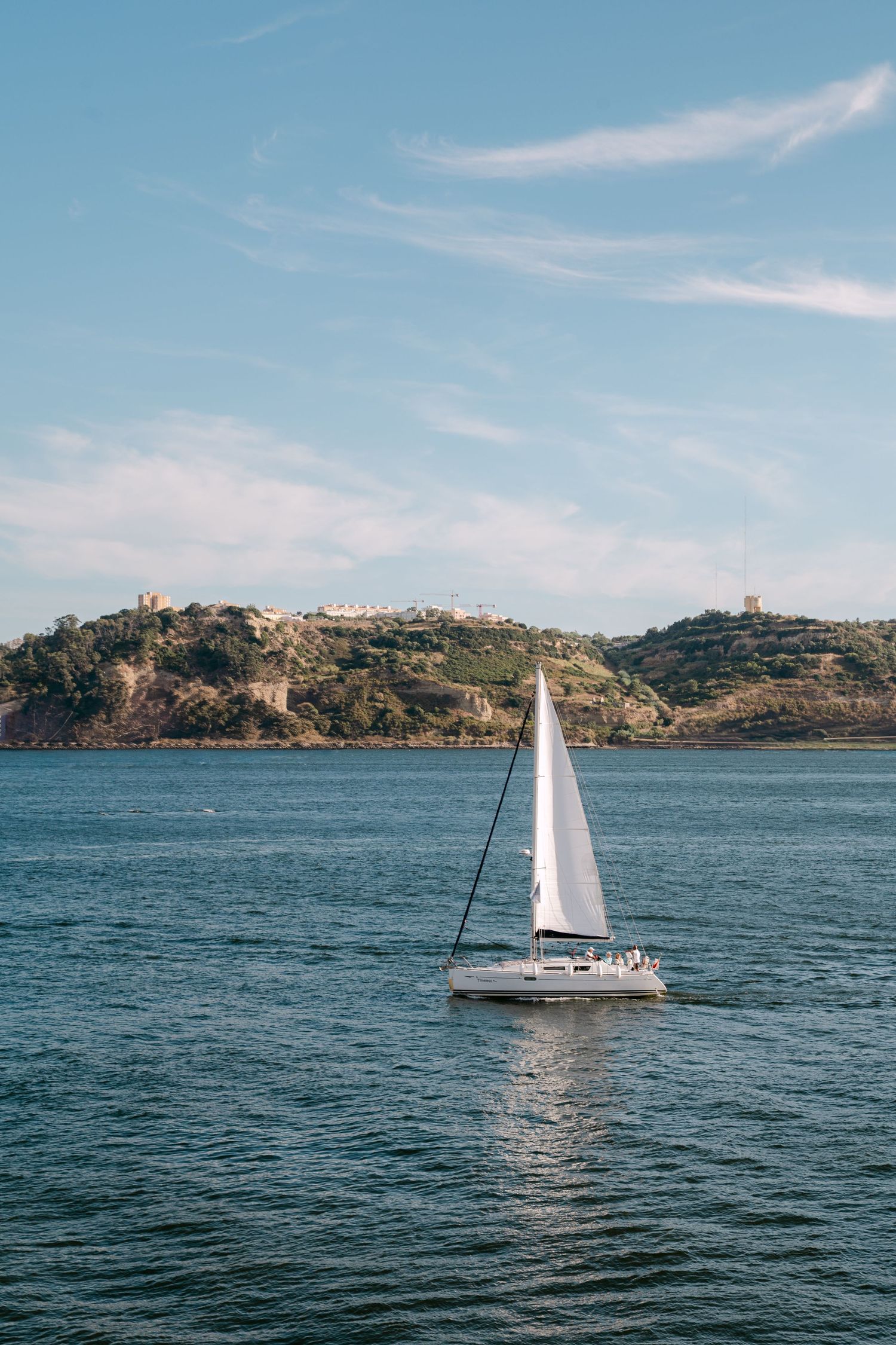 White sailboat gliding across blue waters with coastal hills in the background.
